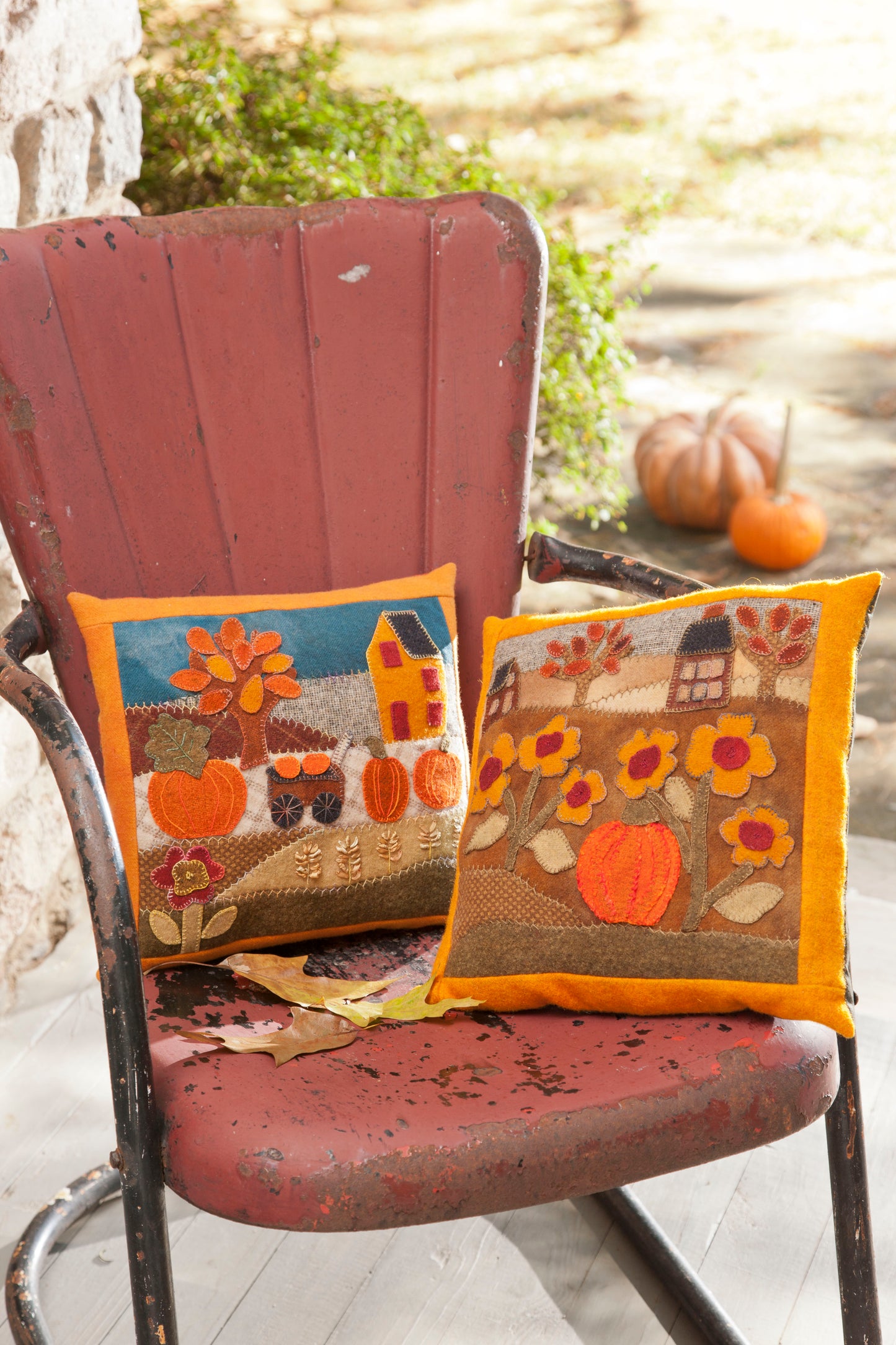A weathered red metal chair on a porch displays two Leisure Arts Cozy Wool Applique Pillows with fall motifs of pumpkins, flowers, and houses. In the background, pumpkins and autumn leaves rest on the ground.
