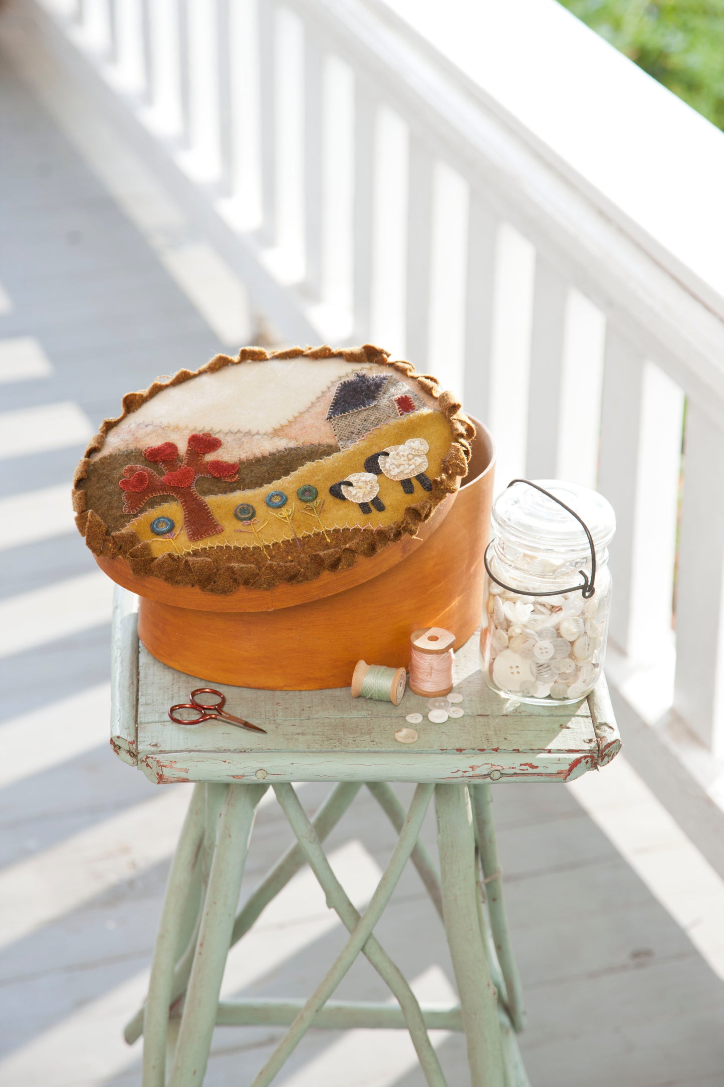 The Cozy Wool Applique Pillows kit by Leisure Arts rests on a vintage green stool beside a glass jar filled with white buttons, thread, and scissors, ready for your next embroidery project on a sunny porch.