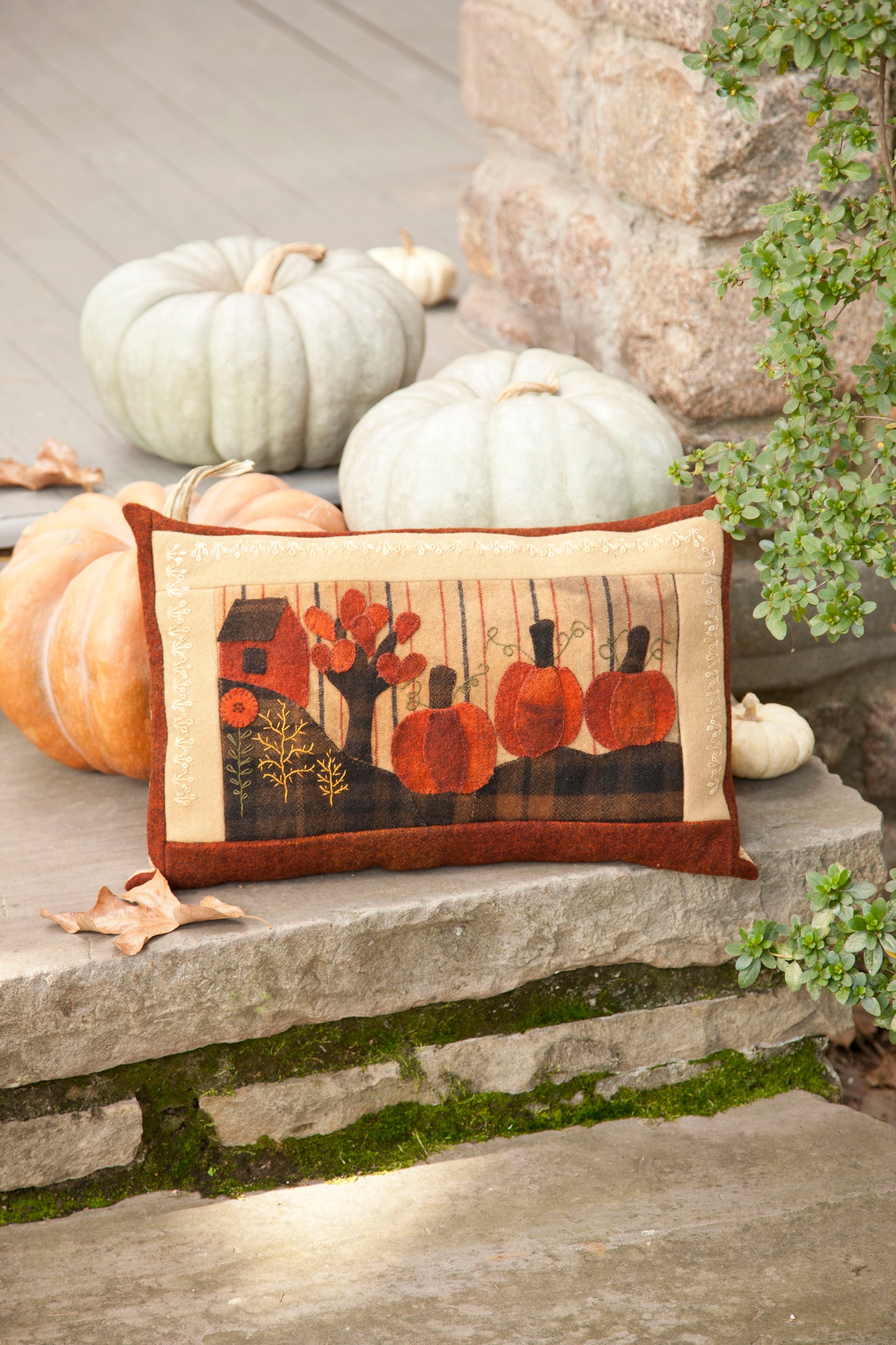 The Cozy Wool Applique Pillow by Leisure Arts, featuring a fall pattern with embroidered pumpkins and flowers, rests on a stone step beside white and orange pumpkins and lush green plants.