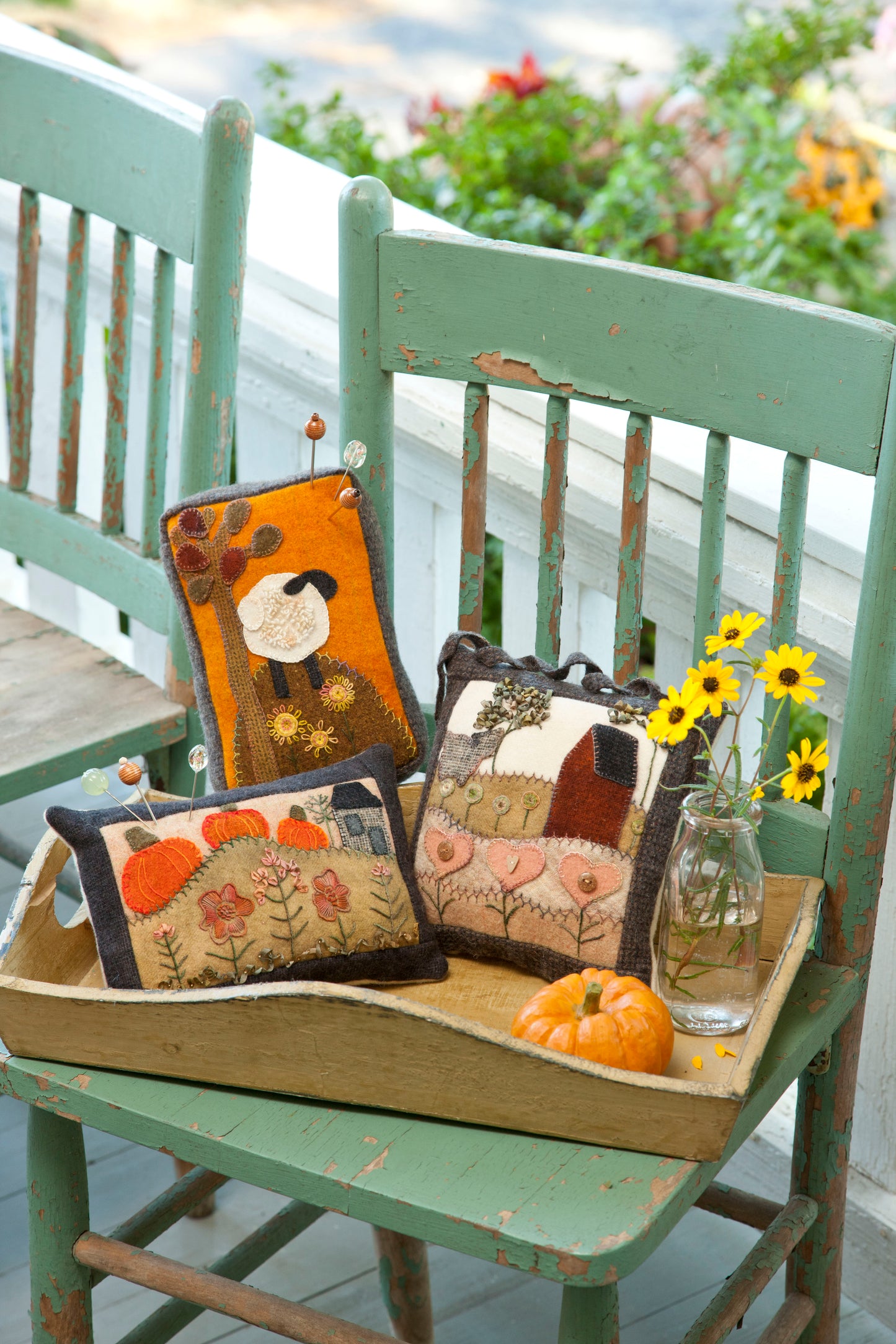 A vintage green chair displays a wooden tray with three Cozy Wool Applique Pillows by Leisure Arts, a small pumpkin, and a glass jar of yellow flowers—perfect autumn décor for your porch. A matching chair stands nearby.