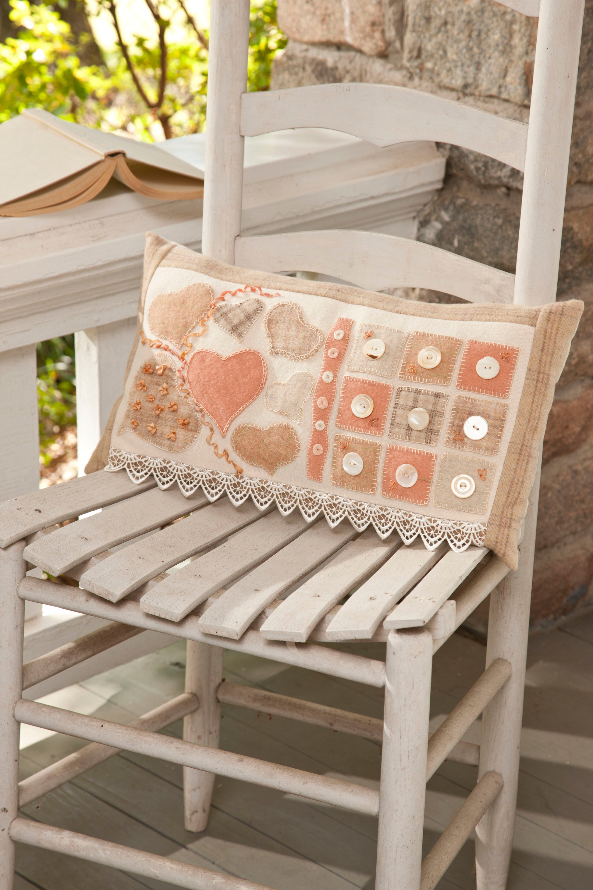 The Cozy Wool Applique Pillows by Leisure Arts, featuring heart and button designs, sit on a rustic white wooden chair on a porch, with an open book visible in the background—perfect for adding charm to your home décor.