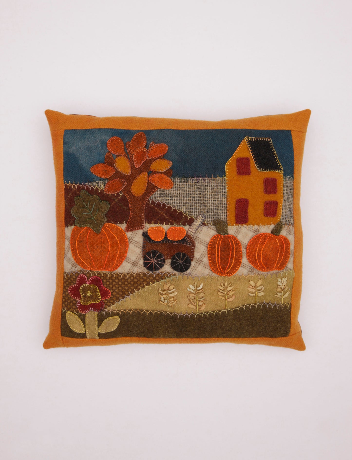 The Cozy Wool Applique Pillows by Leisure Arts feature a charming autumn scene with a tree, pumpkins, house, flowers, and cart on a patchwork backdrop—ideal for home décor or embroidered pillow projects.