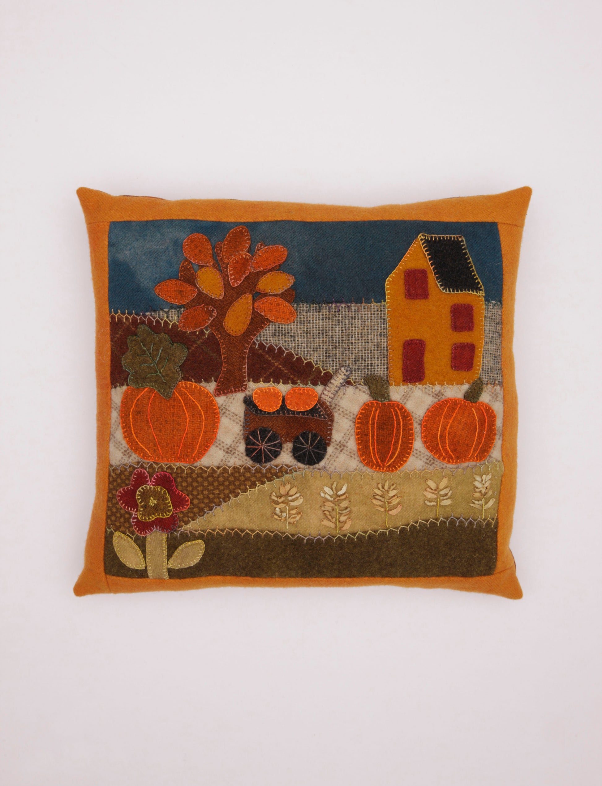 The Cozy Wool Applique Pillows by Leisure Arts feature a charming autumn scene with a tree, pumpkins, house, flowers, and cart on a patchwork backdrop—ideal for home décor or embroidered pillow projects.
