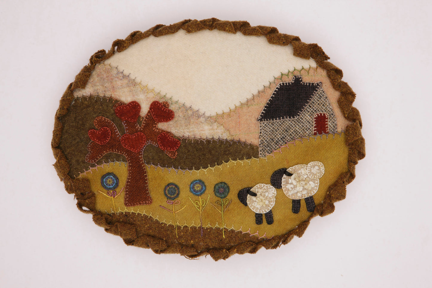 The Cozy Wool Applique Pillows by Leisure Arts feature an oval fabric patch with a house, hills, a red tree, blue flowers, and sheep—ideal for adding rural charm to home décor or embroidery pillow projects.