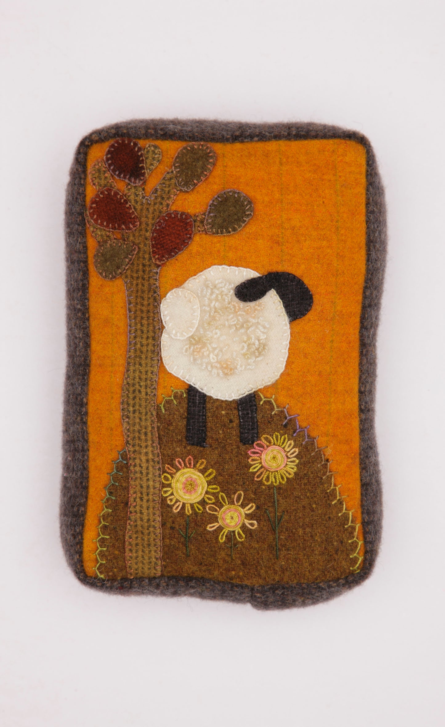 The Cozy Wool Applique Pillows by Leisure Arts feature a rectangular felt patch with a white sheep under a tree, yellow flowers, and an orange background—perfect for home décor or wool embroidery projects.