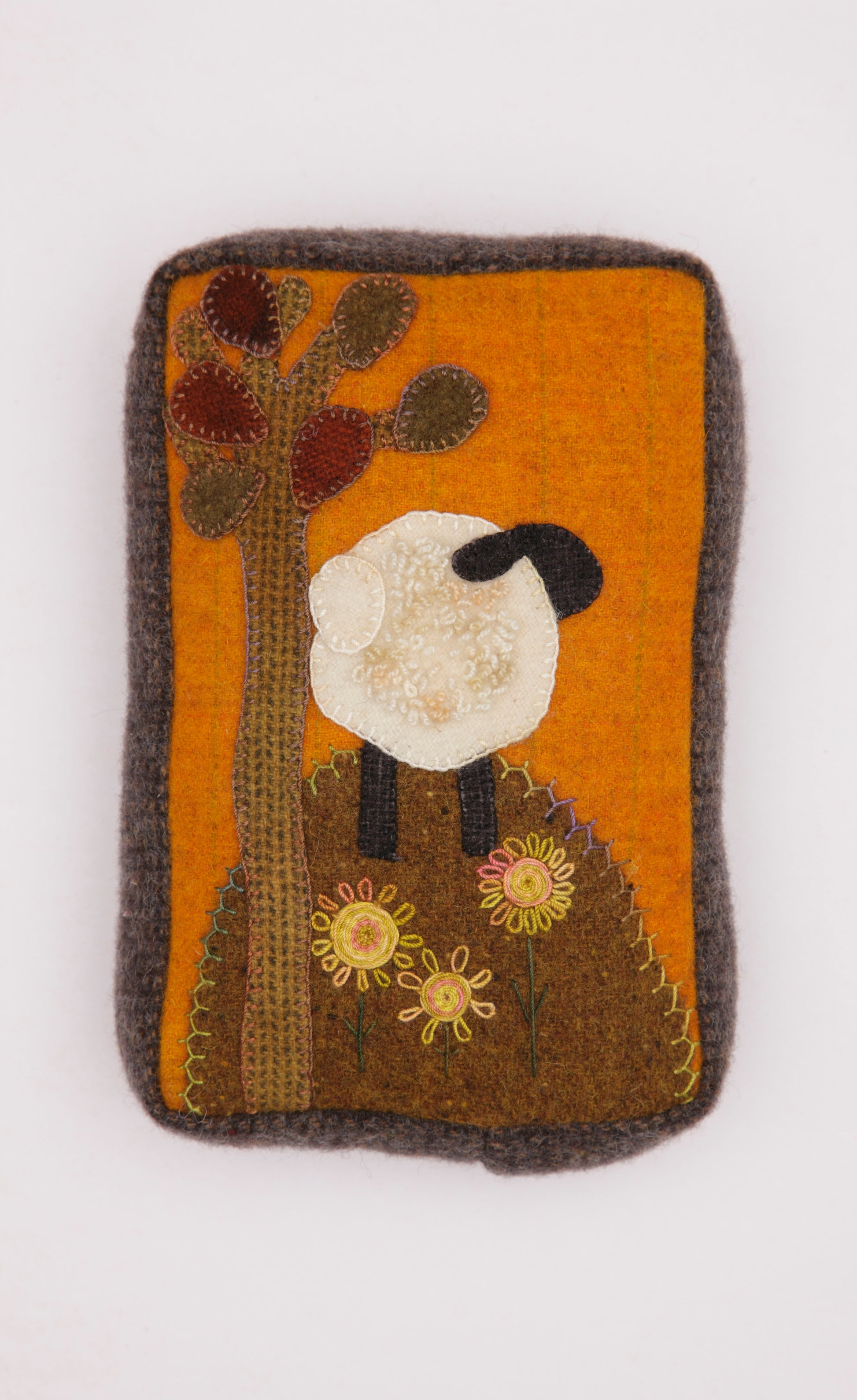 The Cozy Wool Applique Pillows by Leisure Arts feature a rectangular felt patch with a white sheep under a tree, yellow flowers, and an orange background—perfect for home décor or wool embroidery projects.