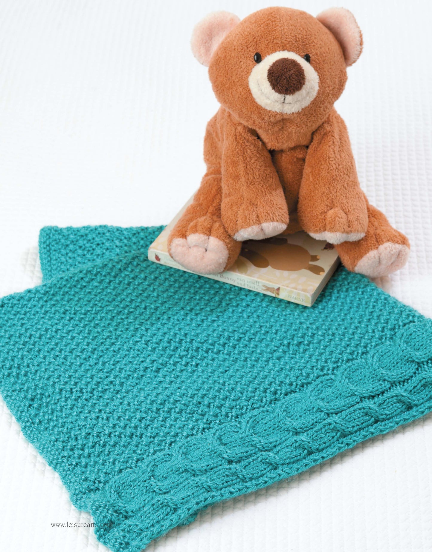 A brown plush teddy bear sits atop the closed "Take the Fear Out of Cables Knitting" book by Leisure Arts, on a textured teal knitted blanket with a decorative border, all arranged on a white surface—ideal inspiration for beginner knitters.