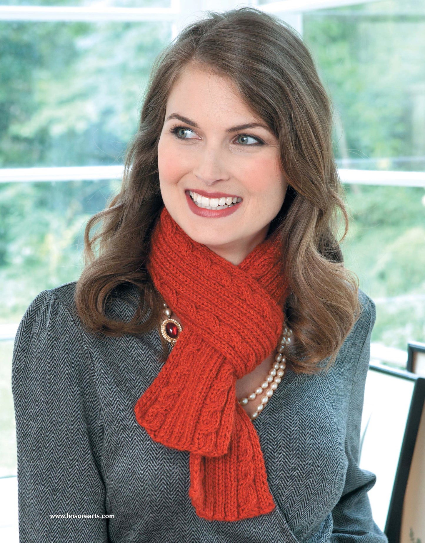 A smiling woman with wavy brown hair wears a textured red knit scarf from "Take the Fear Out of Cables Knitting" by Leisure Arts, paired with a grey patterned jacket and pearl necklace. Large windows and greenery are visible indoors behind her.