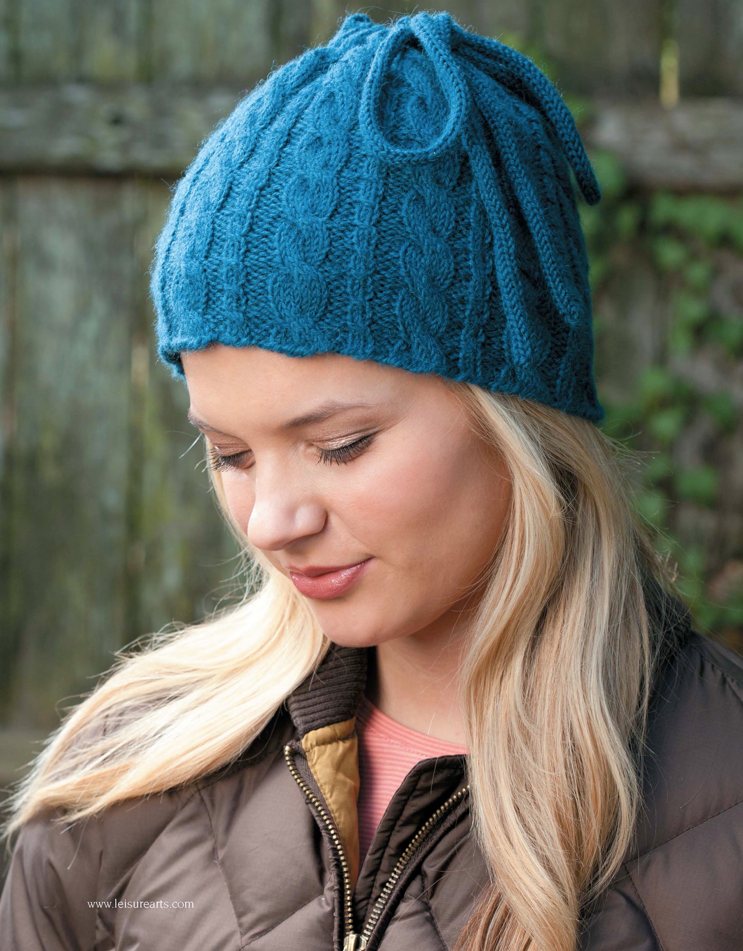 A woman with long blonde hair smiles softly while wearing a teal cable-knit hat made with the “Take the Fear Out of Cables Knitting” pattern by Leisure Arts. She stands in front of a wooden fence, dressed in a brown jacket.
