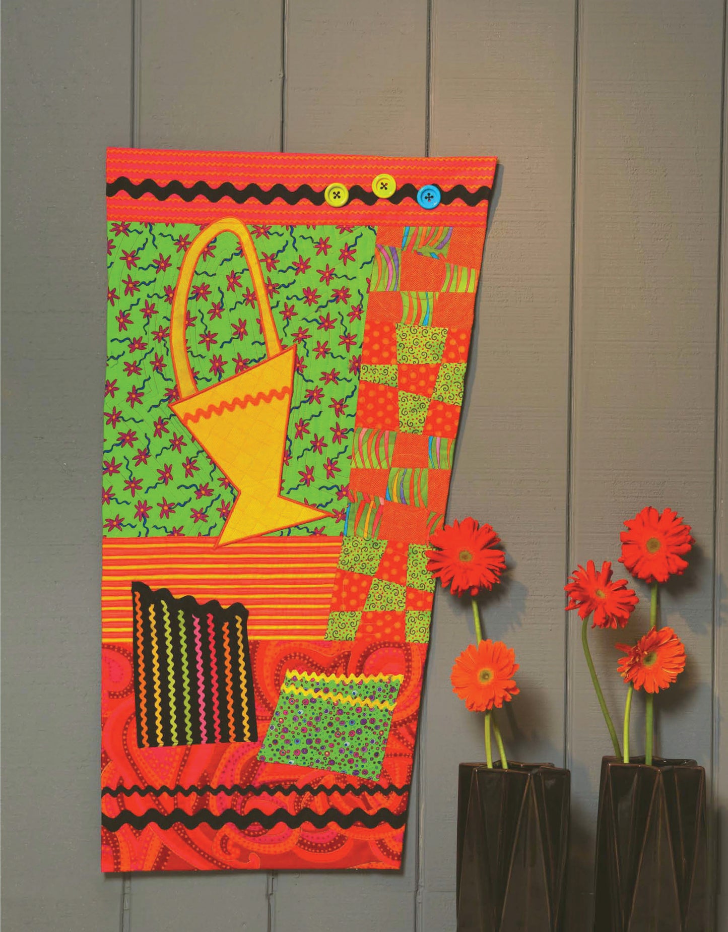 The “Quilt Blocks Go Wild!” quilt by Leisure Arts hangs on a gray wall, displaying vibrant red, green, yellow, and blue shapes with buttons. A yellow basket and two black vases of orange-red gerbera daisies add lively accents—ideal for beginner quilters.