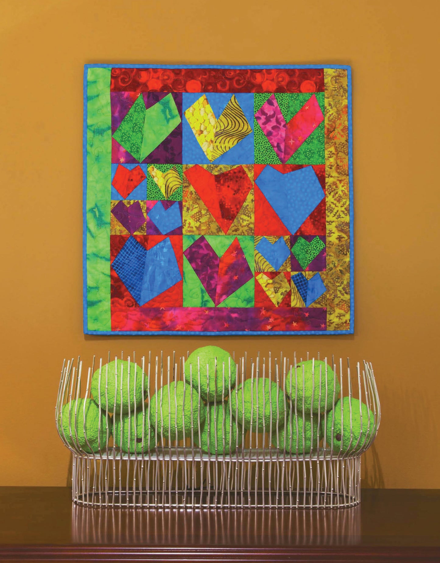 Leisure Arts' Quilt Blocks Go Wild! wall quilt, featuring heart shapes and easy patterns for beginners, hangs above a white wire basket holding bright green tennis balls, set against an orange wall.