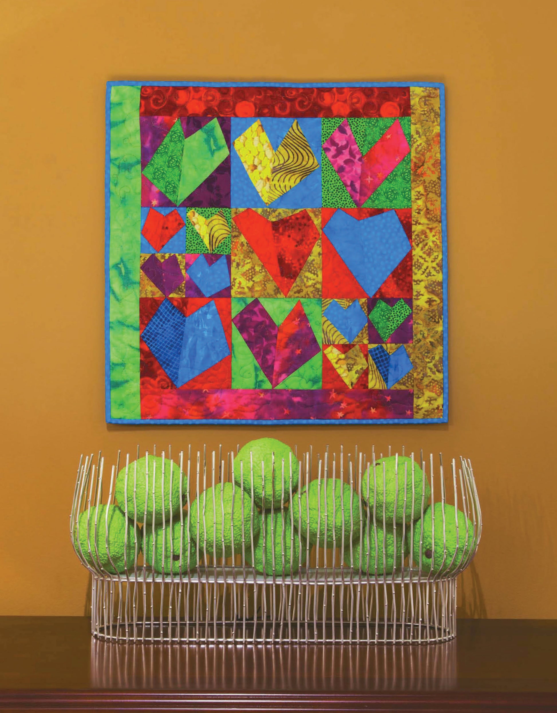 Leisure Arts' Quilt Blocks Go Wild! wall quilt, featuring heart shapes and easy patterns for beginners, hangs above a white wire basket holding bright green tennis balls, set against an orange wall.