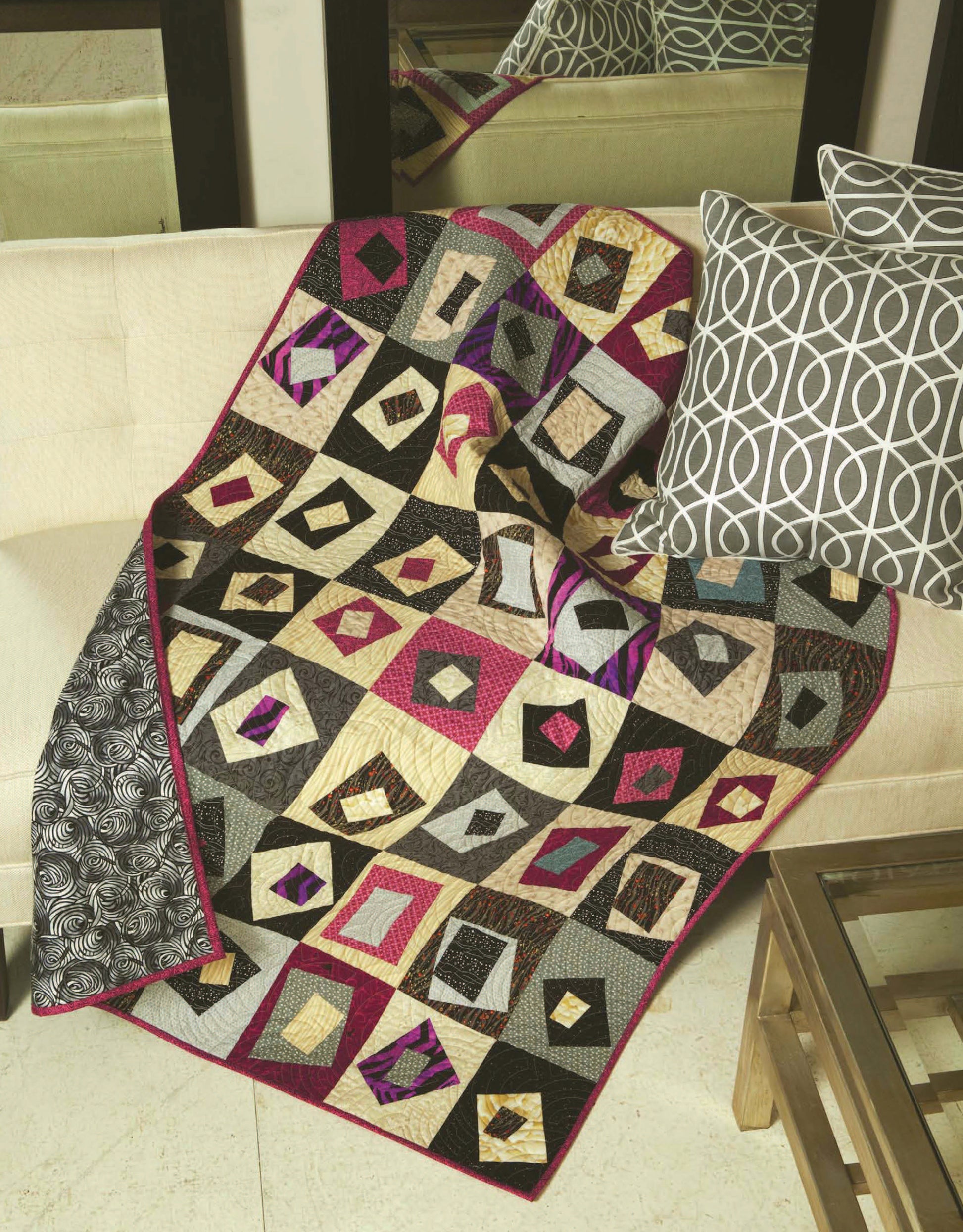 The Quilt Blocks Go Wild! patchwork quilt by Leisure Arts, with bold patterns, is draped over a beige couch beside two white and gray abstract pillows. A glass coffee table sits in front.