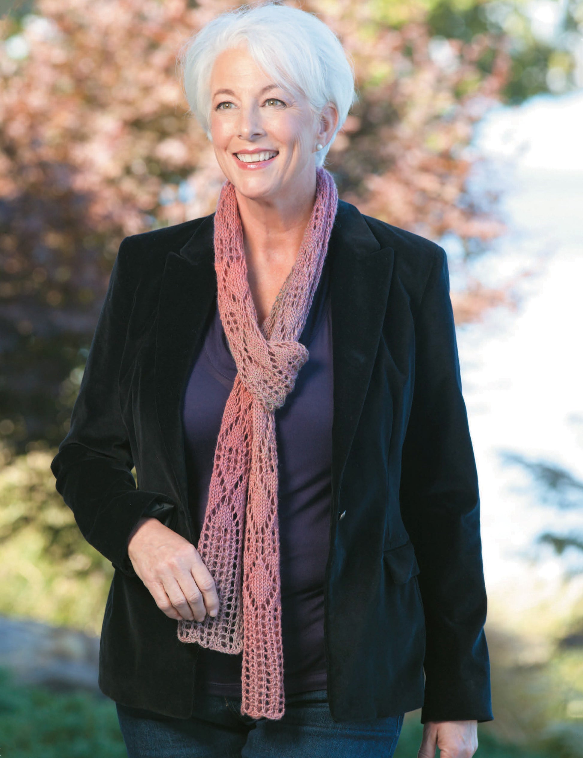 Smiling woman with short white hair wears a black blazer, dark top, and her favorite accessory—a pink lace scarf made from *Knitting for a Cure* by Leisure Arts—while standing outdoors with trees and water behind her.