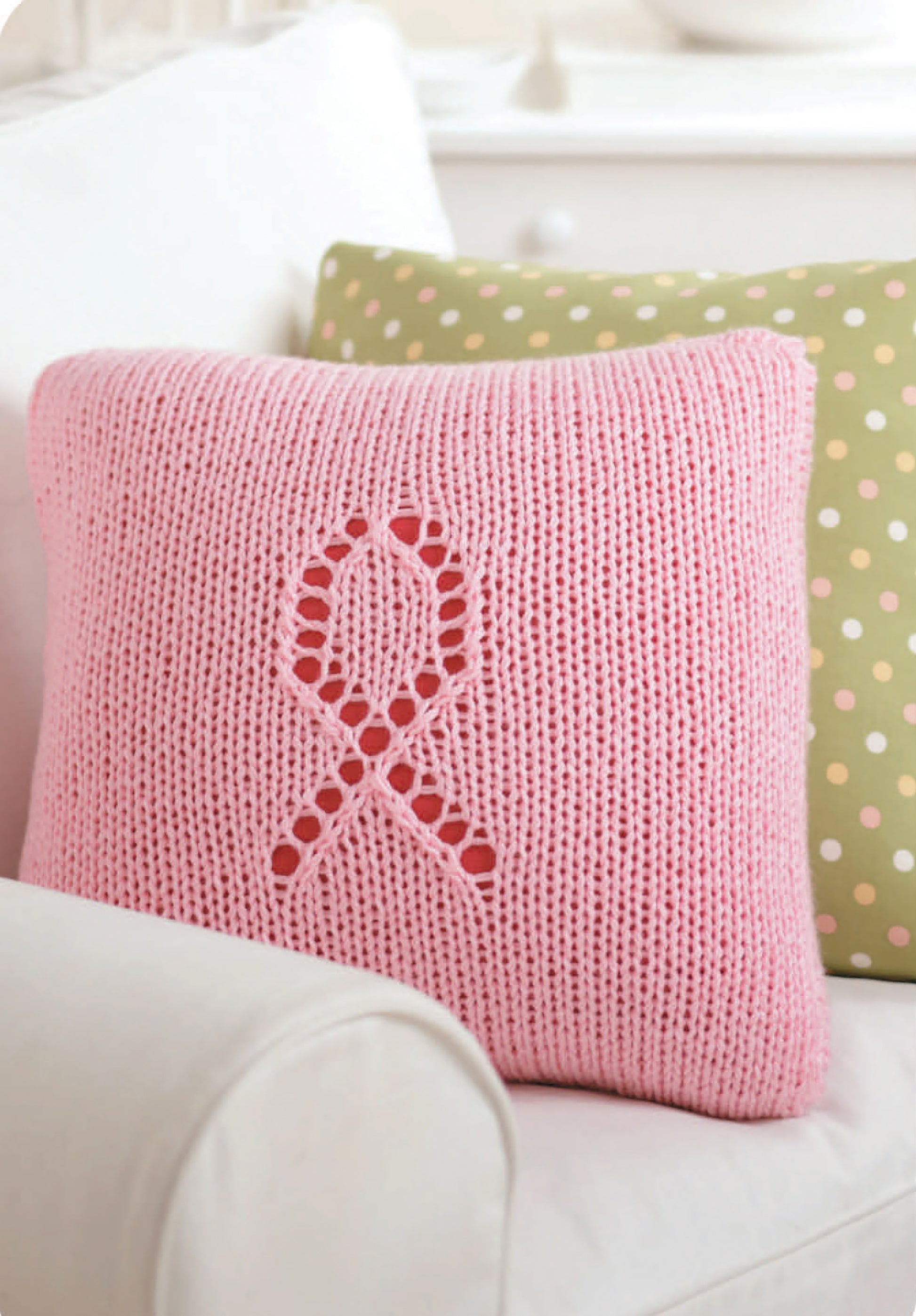 The "Knitting for a Cure" pillow by Leisure Arts, featuring a breast cancer ribbon design, sits on a white couch to show support, while a green polka dot cushion adds charm in the background.