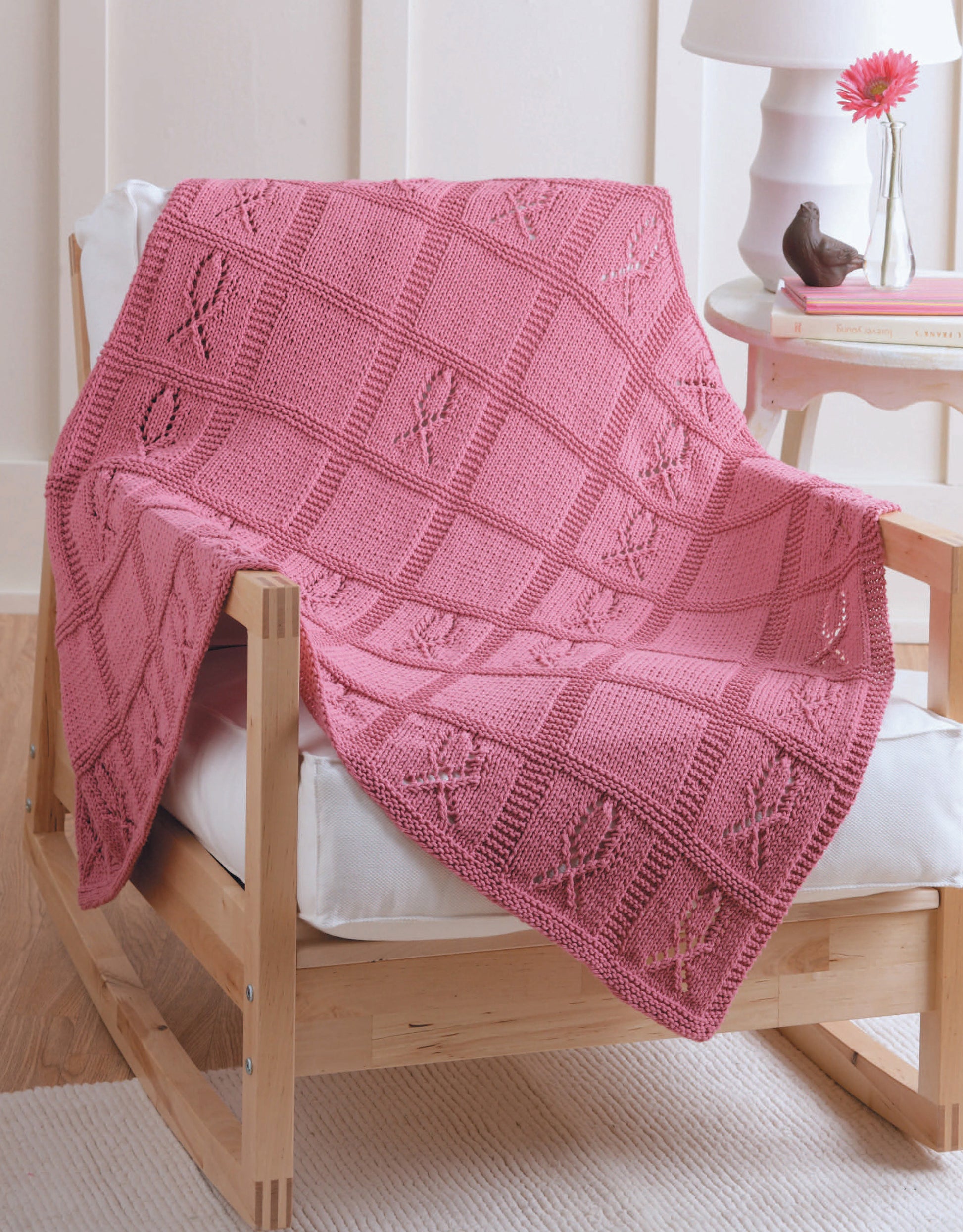 The Knitting for a Cure blanket by Leisure Arts, featuring pink awareness ribbons for breast cancer support, is draped over a light wood chair in a cozy room with books, a lamp, and flowers.