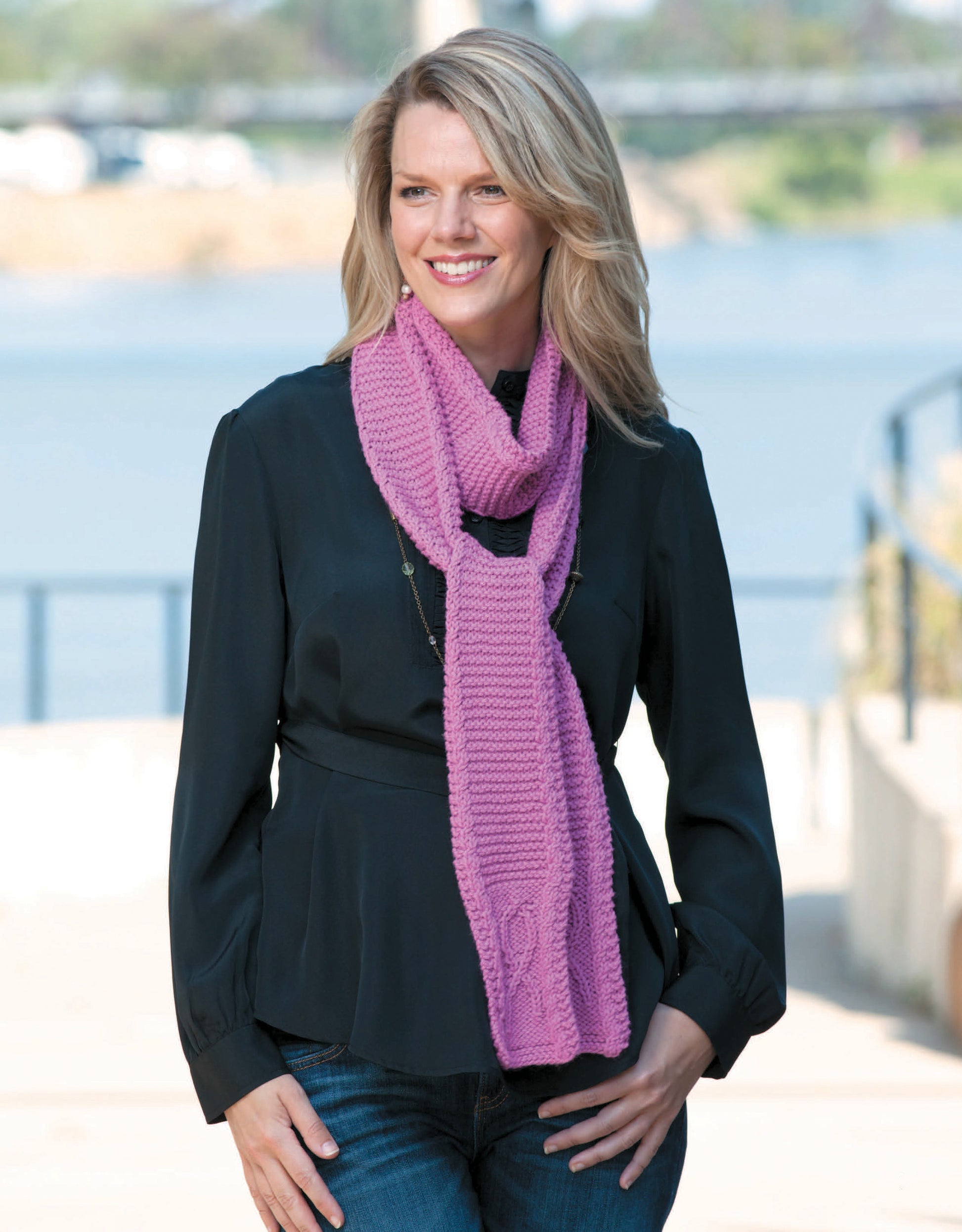 A woman with blonde hair, in a black long-sleeve blouse and blue jeans, smiles by the water wearing Leisure Arts' "Knitting for a Cure" pink scarf—a stylish accessory wrapped around her neck.