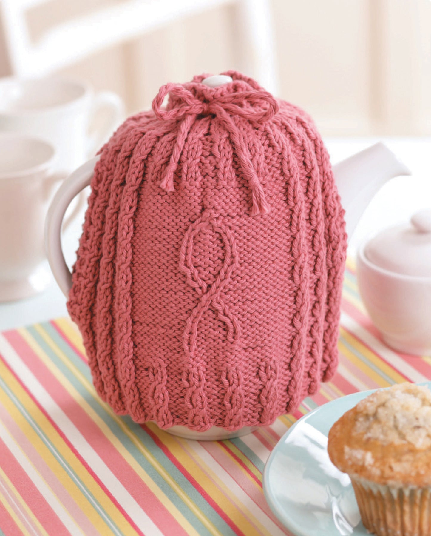 The "Knitting for a Cure" teapot cozy by Leisure Arts, featuring pink cable patterns, sits on a striped placemat with a muffin, cups, and sugar bowl—adding charm while subtly supporting breast cancer awareness.