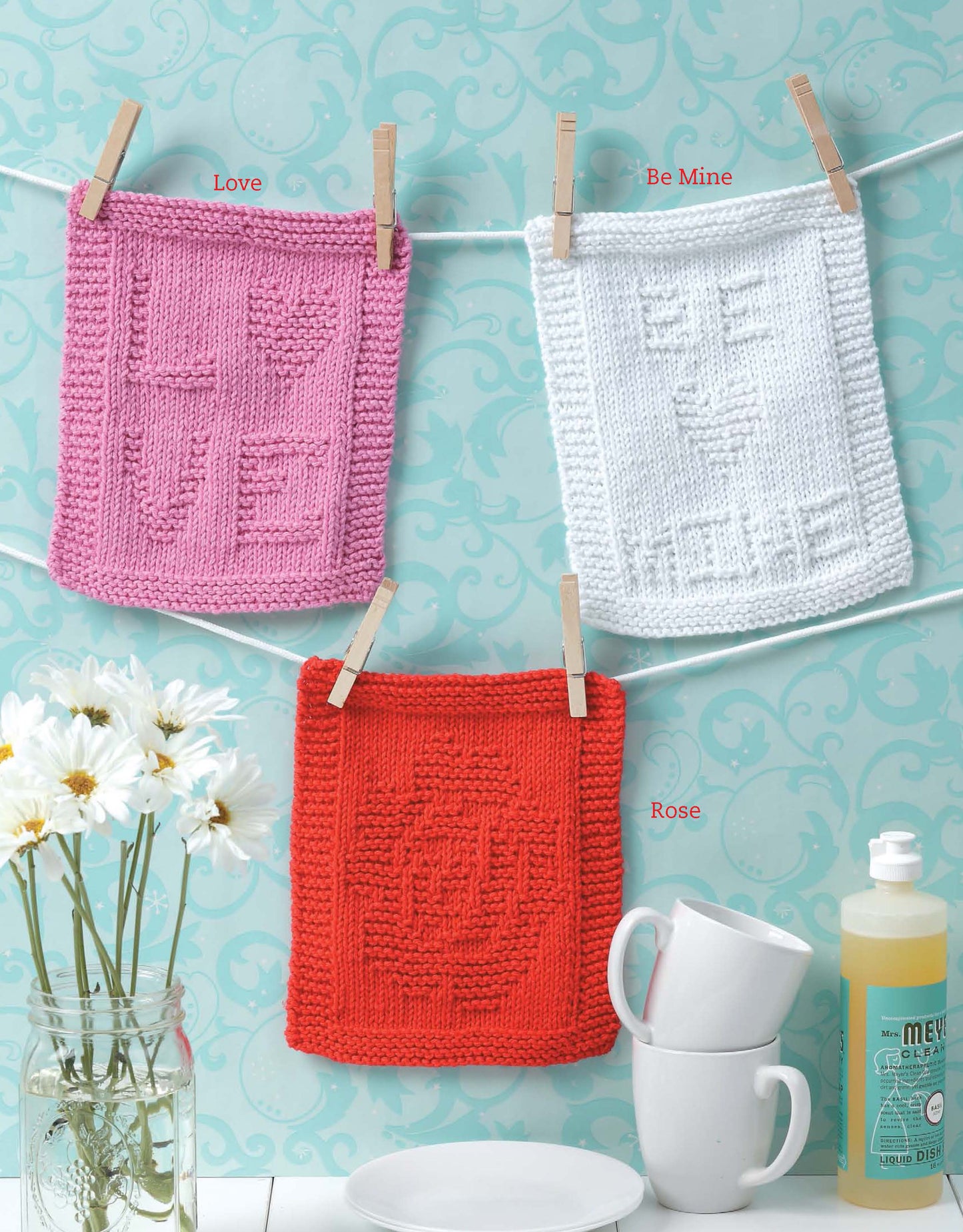 Three dishcloths in pink (LOVE), white (BE MINE), and red hang on a string with clothespins. The Leisure Arts "Dishcloths for Special Days" digital download offers easy knitting projects—great for beginners or as quick, charming gifts.