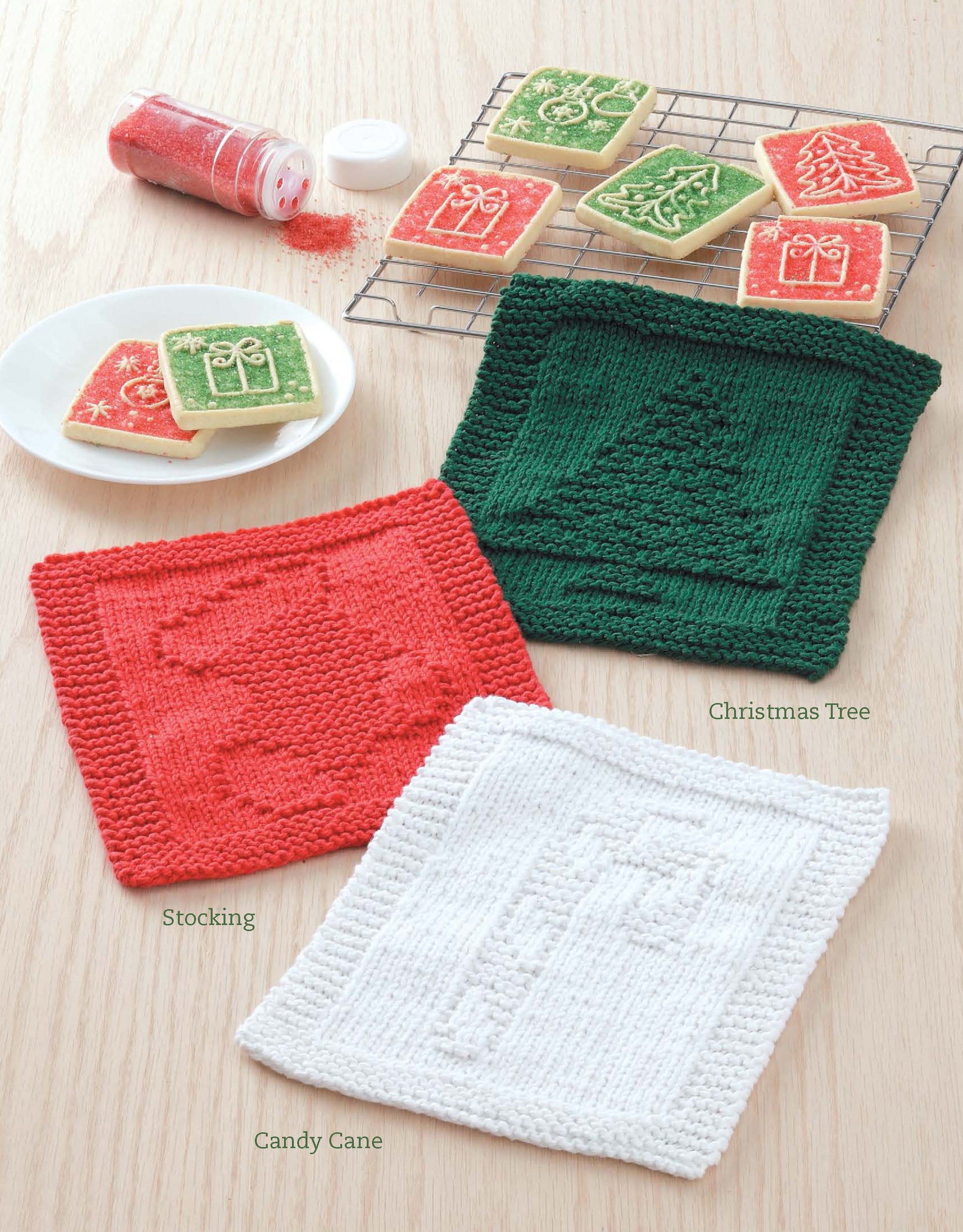 The Dishcloths for Special Days Digital Download by Leisure Arts features festive knit washcloths—red with stocking, green with Christmas tree, white with candy cane—ideal as gifts or beginner projects, shown beside decorated holiday cookies.