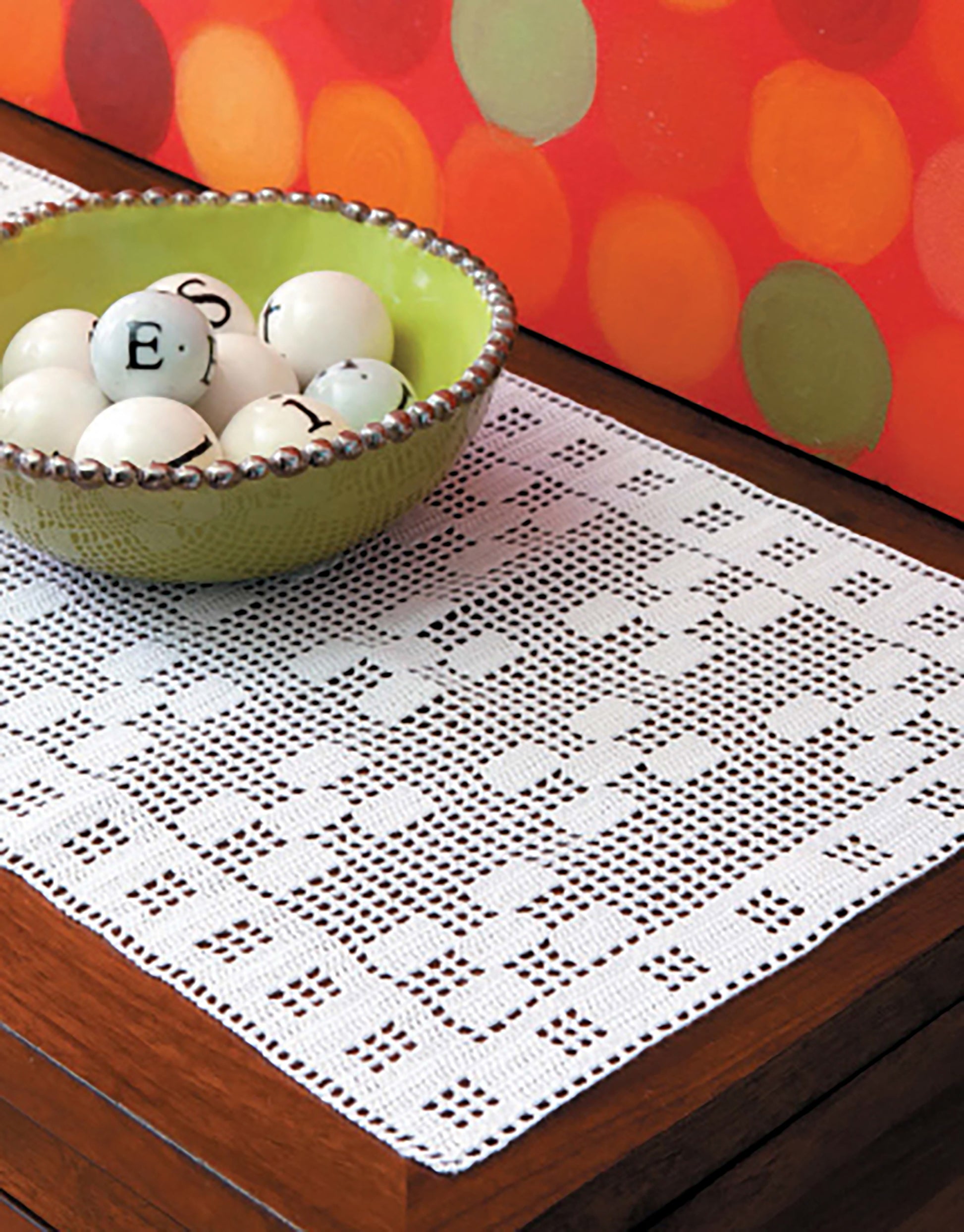 The "Filet Table Runners Digital Download" by Leisure Arts features a white geometric crochet runner on wood, topped with a green bowl of white lettered balls, set against a bold red and orange polka dot backdrop.