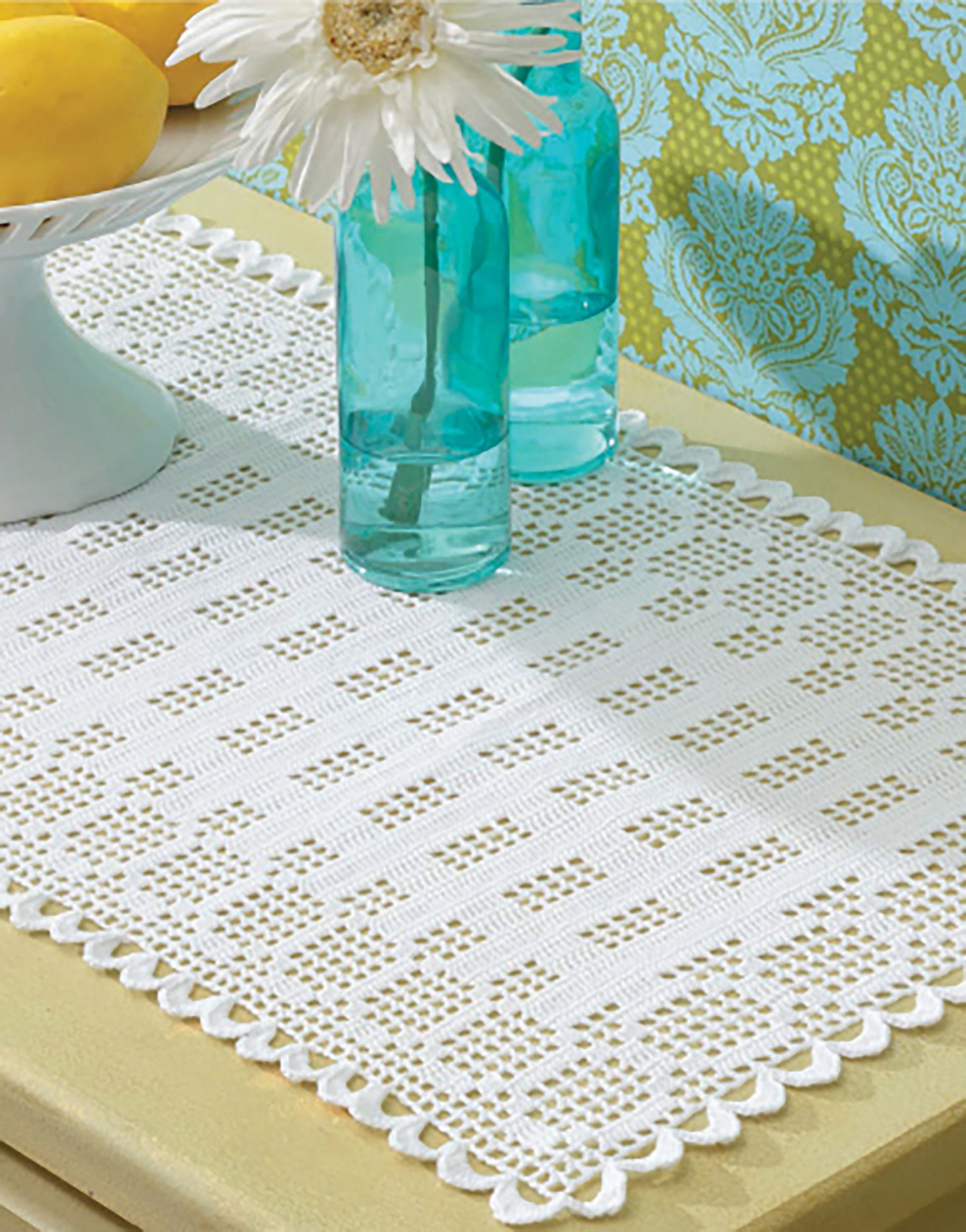 The Filet Table Runners Digital Download by Leisure Arts features a white lace runner with scalloped edges, styled on a table with blue glass bottles, lemons in a white dish, and floral wallpaper—a great idea for beginner crochet projects.