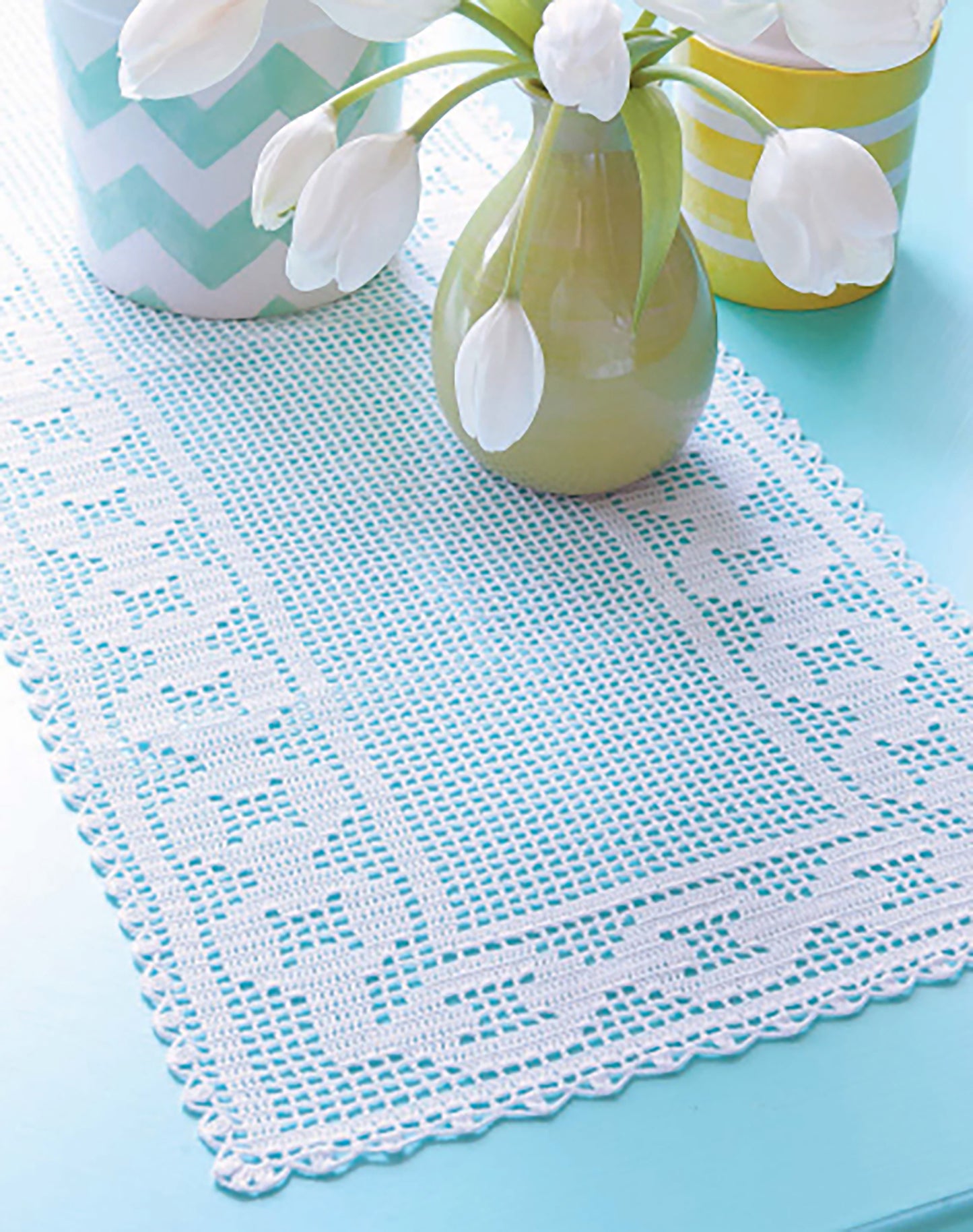 The Filet Table Runners Digital Download by Leisure Arts features a white crochet runner with a geometric border, displayed on a light blue table beside a green vase of white tulips and a striped yellow-green container.