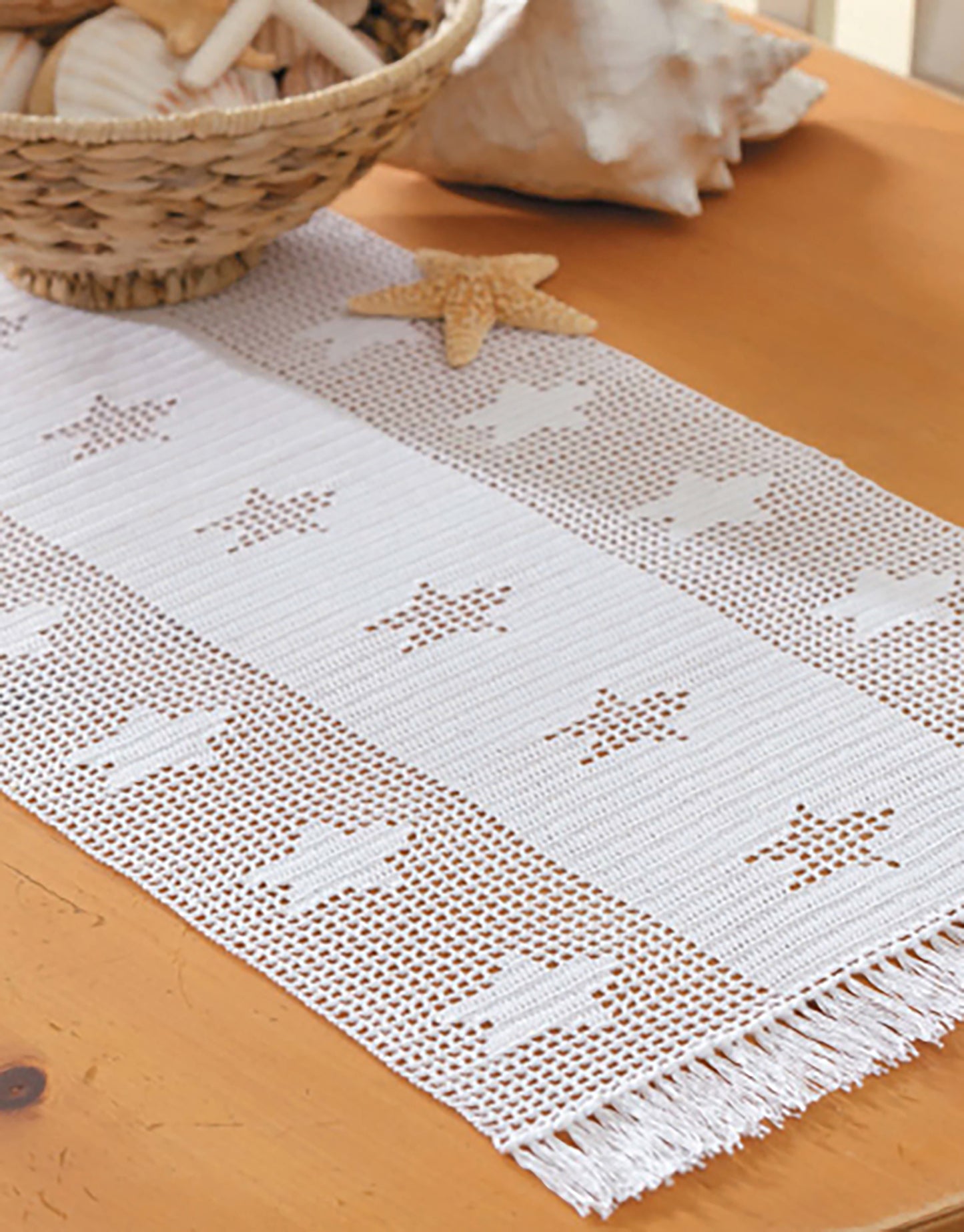 The Leisure Arts Filet Table Runners Digital Download features a white crochet lace runner with star and shell motifs and fringed edges, perfect for beginners. Styled on wood with seashells, it beautifully complements a beach-themed decor.
