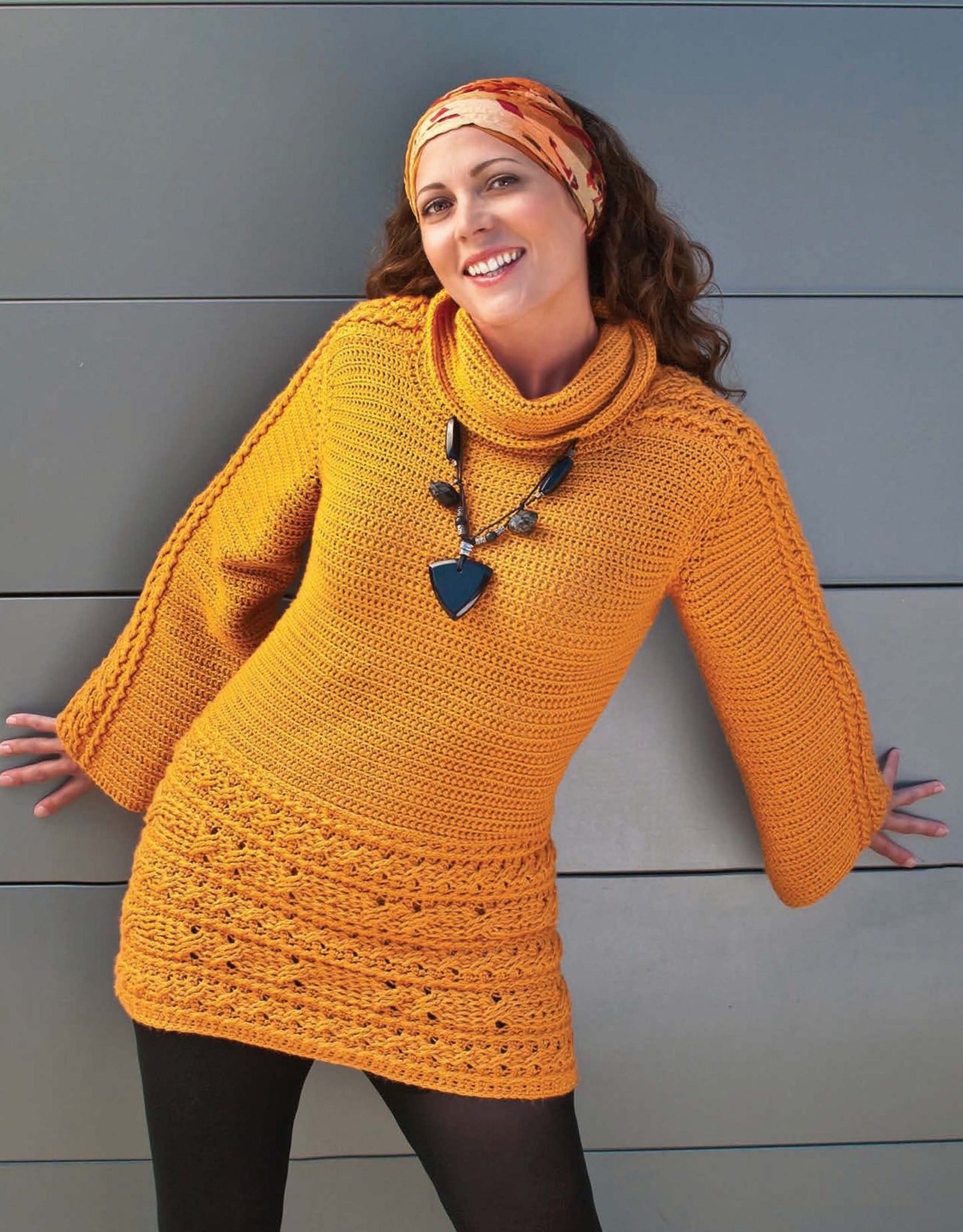 A woman models the Crochet Urban Edge Fashion sweater dress by Leisure Arts, pairing it with a patterned headband and a chunky black necklace as she poses against a gray wall, arms slightly outstretched.