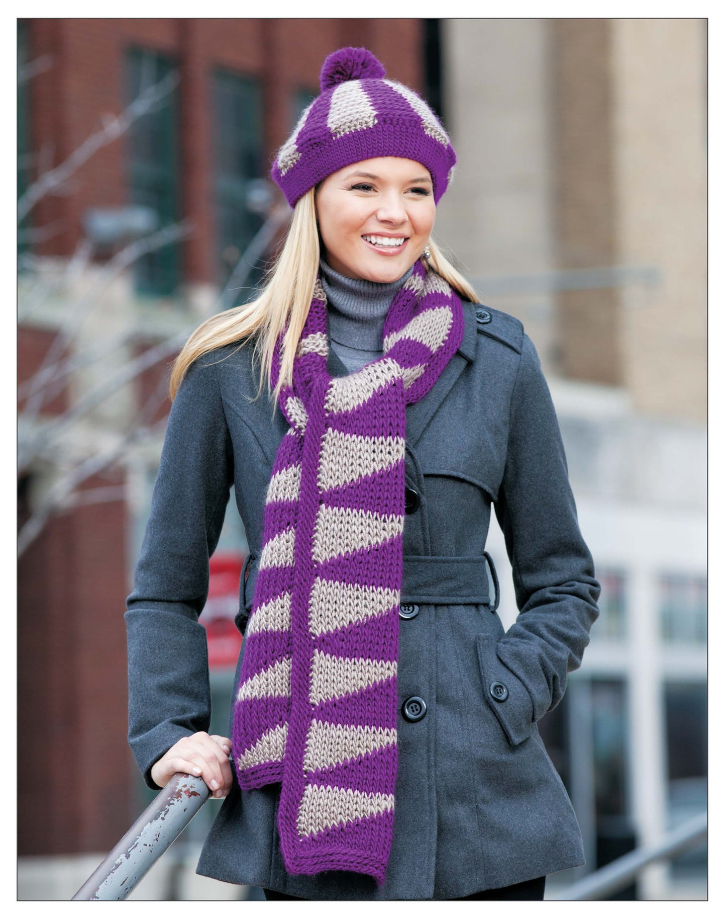 A smiling person wears a grey coat and a purple and beige striped scarf made with the Leisure Arts Short Row Tunisian Stitch Crochet, plus a matching purple pom-pom hat, while standing outdoors in a city.