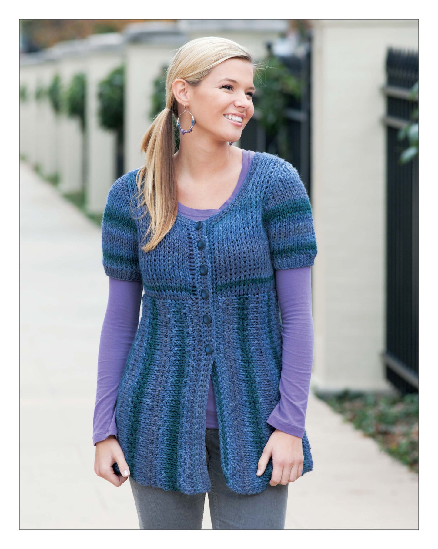 A smiling woman outdoors wears the Leisure Arts Short Row Tunisian Stitch Crochet short-sleeve blue and purple cardigan over a long-sleeve purple shirt with gray pants. She has blond hair in a low side ponytail and large hoop earrings.