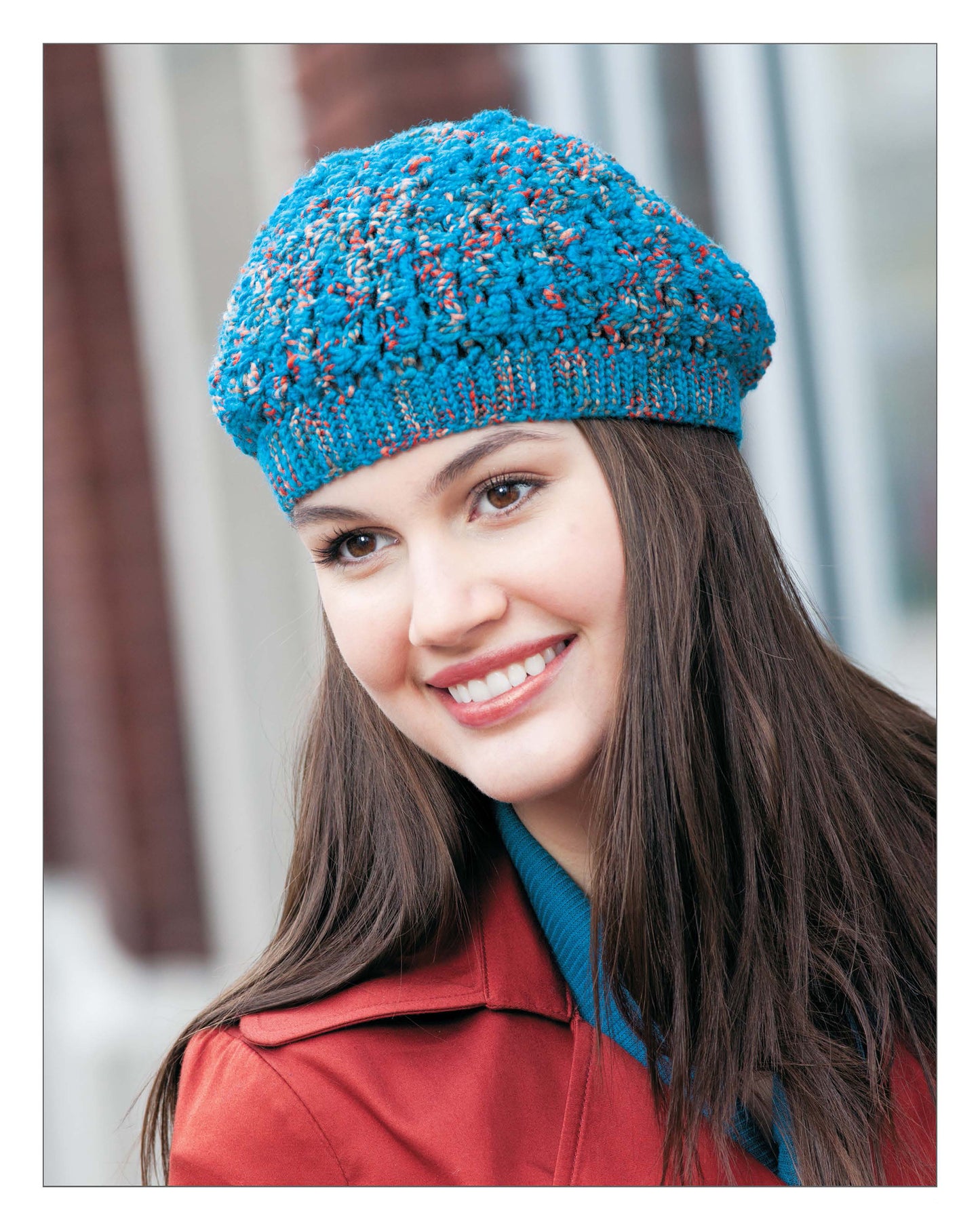 A young woman with long brown hair smiles, wearing the Leisure Arts Short Row Tunisian Stitch Crochet blue beret and a red coat over a teal top. The softly blurred background suggests she’s outdoors.