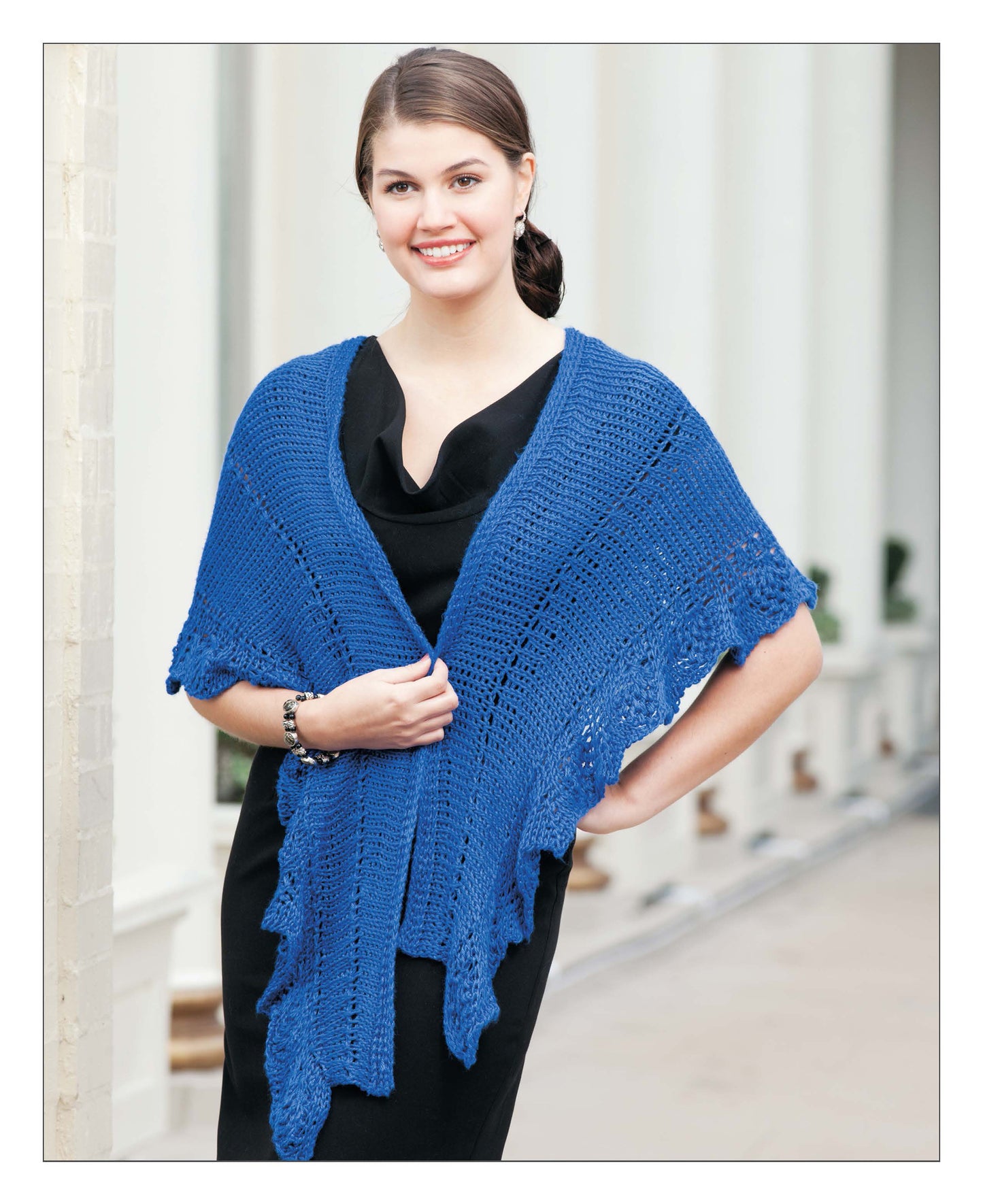 A smiling woman in a black dress stands outside, draped in the blue "Short Row Tunisian Stitch Crochet" shawl by Leisure Arts. She holds the crochet shawl with both hands, her brown hair pulled back. White columns can be seen in the background.