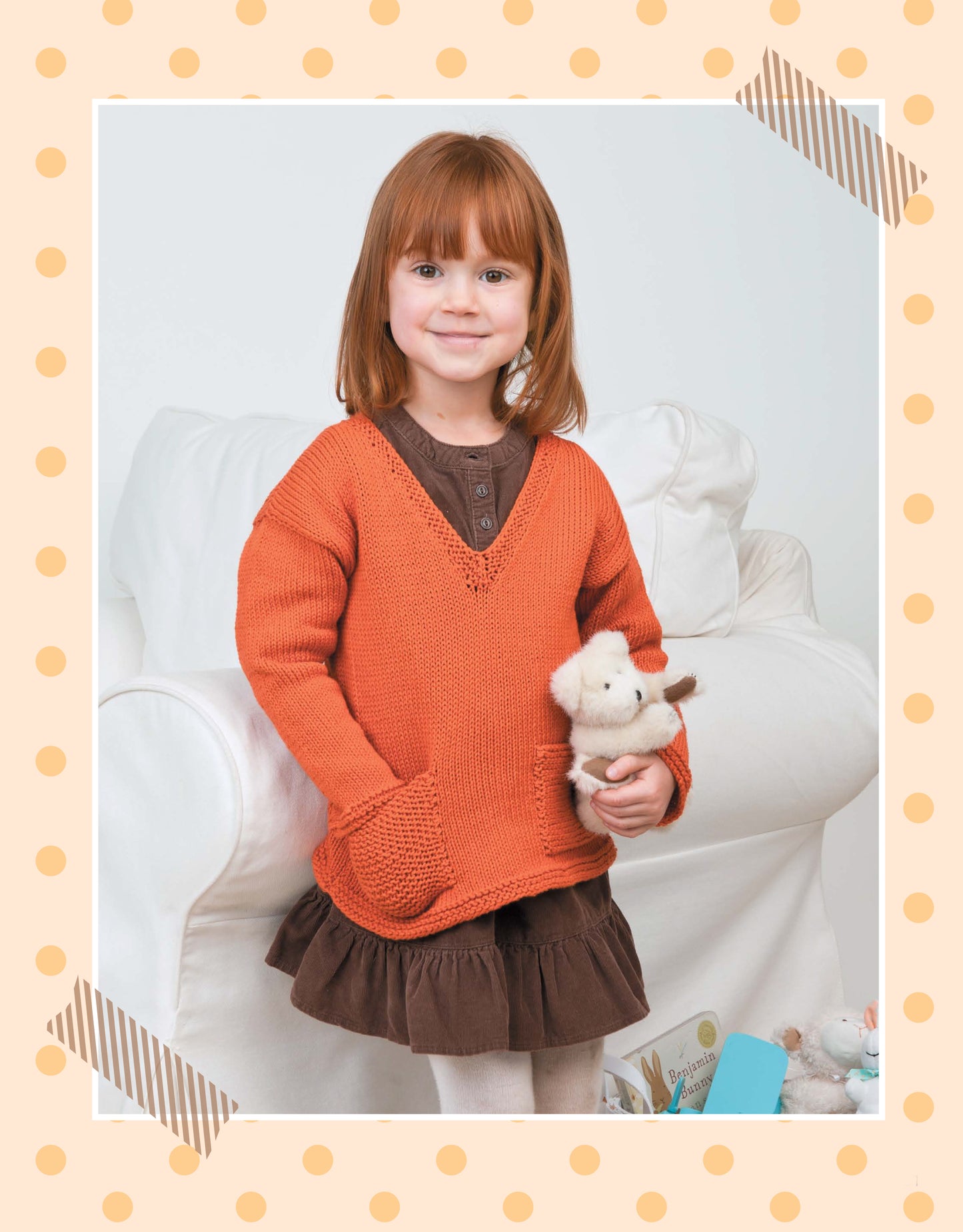 A young red-haired girl in an orange sweater made with the Pick the Pieces Knitting pattern by Leisure Arts stands before a white armchair, smiling and holding a stuffed animal. A beige polka dot border frames the image.