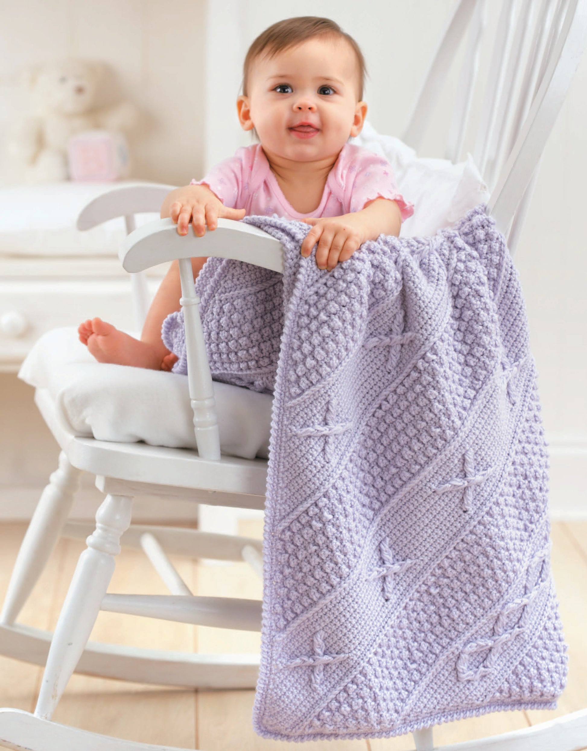 A baby in a pink outfit smiles while sitting in a white rocking chair, holding the Leisure Arts Baby's Diagonal Aran Afghans, a light purple crochet blanket draped over the side, in a cozy, softly lit room.