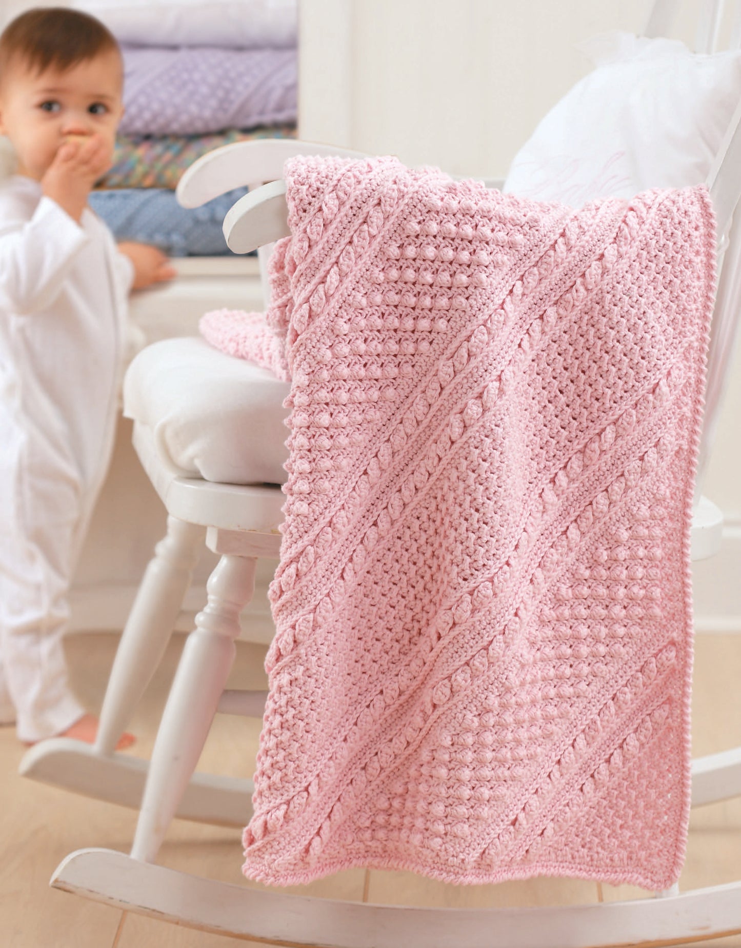 A baby in white pajamas stands near a soft pink crochet blanket from Leisure Arts’ Baby's Diagonal Aran Afghans, which drapes over a white rocking chair—ideal inspiration for those looking for baby crochet patterns.
