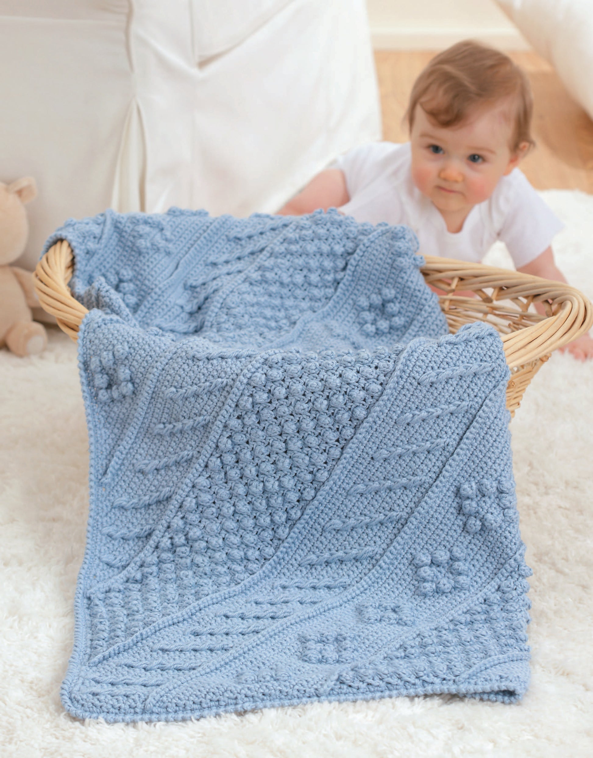 A baby in a white outfit crawls beside a wicker basket draped with Leisure Arts' Baby's Diagonal Aran Afghans—a light blue knit blanket featuring textured patterns and raised flower designs inspired by classic baby crochet styles.