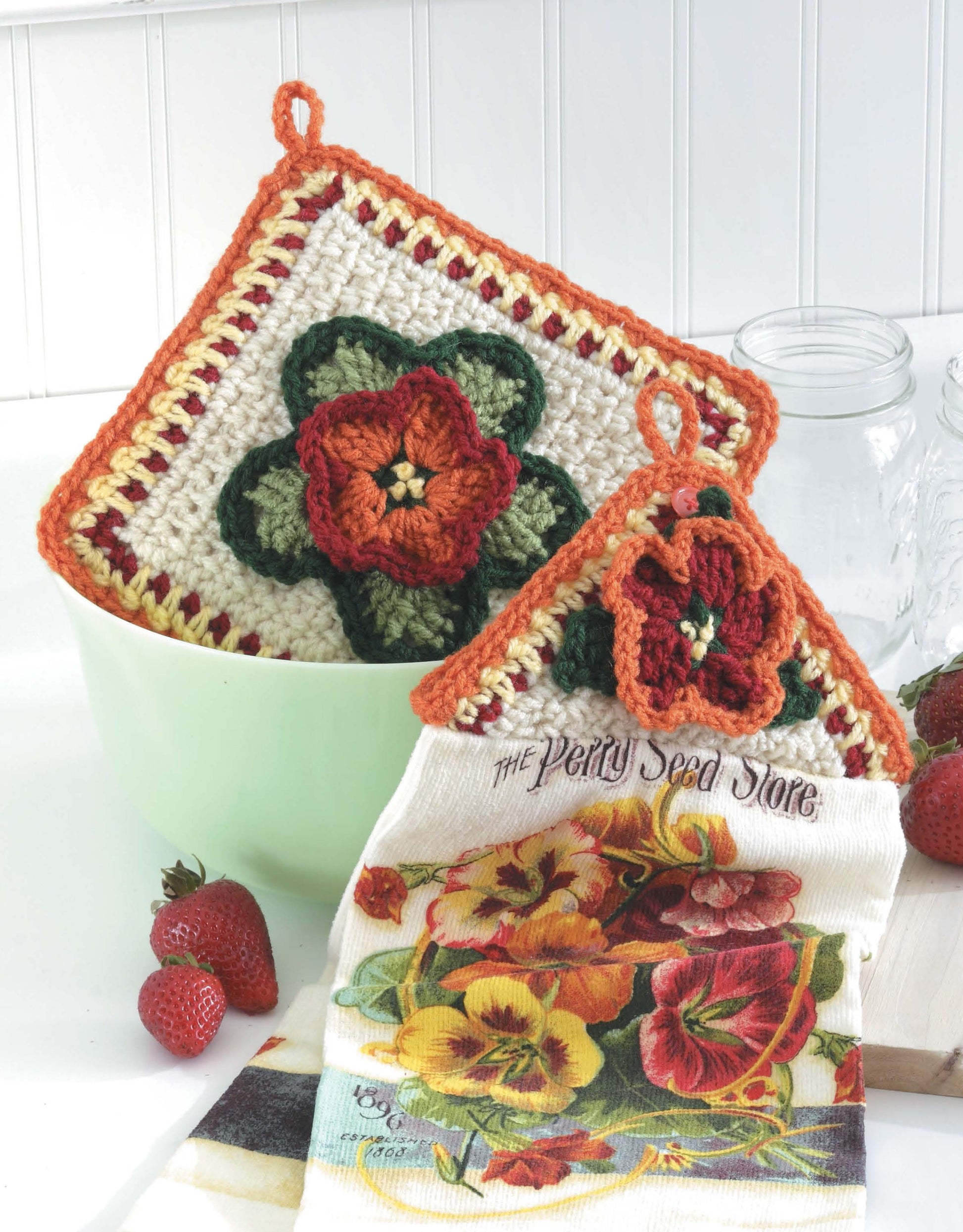 Leisure Arts' Towel Toppers and Potholders Digital Download features colorful crochet flower designs—perfect kitchen decor or thoughtful housewarming gifts. Displayed on a green bowl with strawberries, a floral towel, and glass jars.