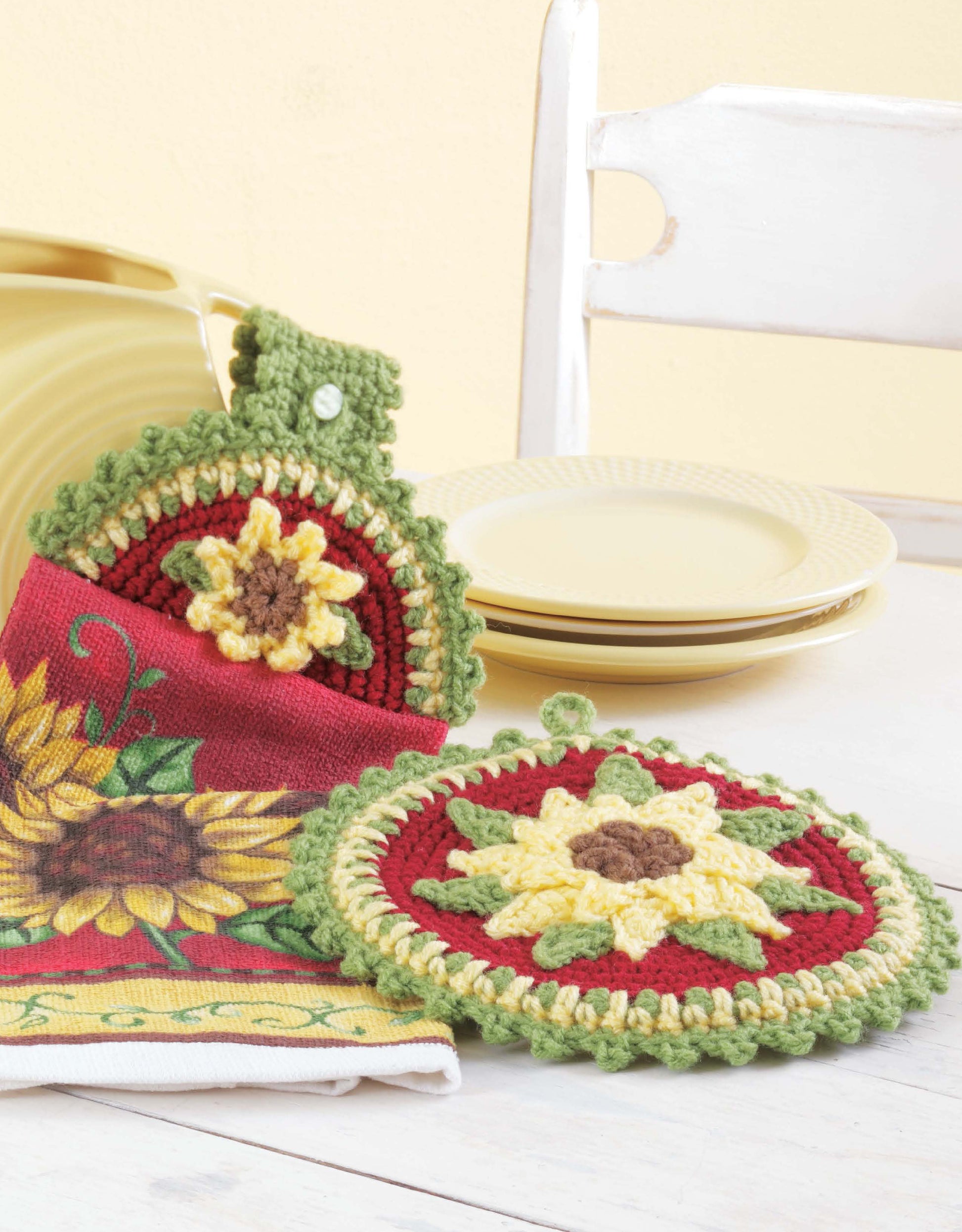 Two Towel Toppers and Potholders, a digital download from Leisure Arts, featuring sunflower designs, are displayed as vibrant kitchen decor alongside a matching towel, yellow plates and bowl, with a white chair in the background.