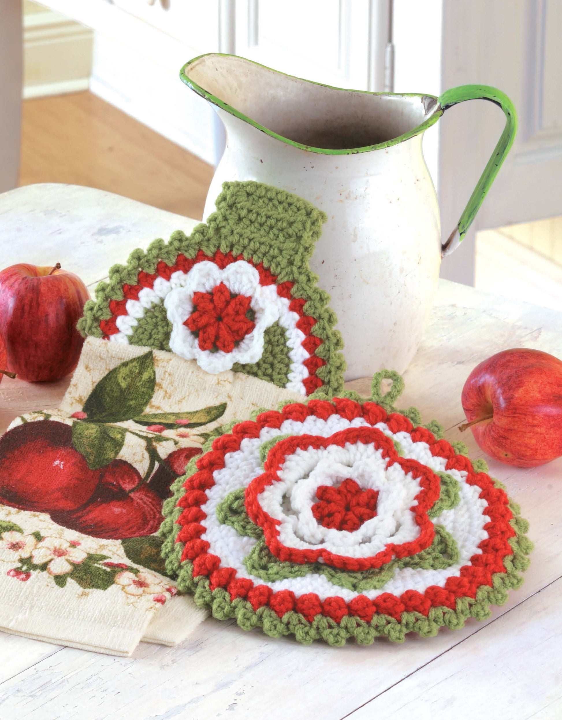 A white pitcher with a green rim sits beside two red apples, apple-themed napkins, and Leisure Arts’ Towel Toppers and Potholders Digital Download—colorful crochet designs that make perfect housewarming gifts.