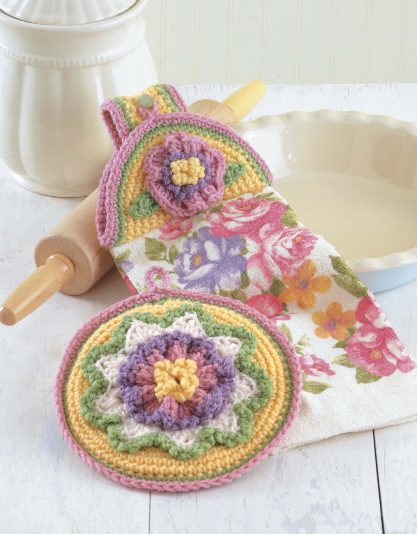 The Leisure Arts "Towel Toppers and Potholders" digital download features pastel, floral crochet designs—ideal for kitchen decor or housewarming gifts—displayed beside a dish towel, rolling pin, pie dish, and white canister.