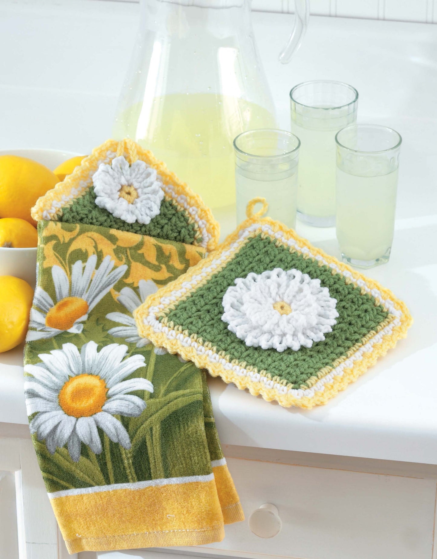 A pitcher and two glasses of lemonade sit beside a bowl of lemons, a floral towel, and a daisy potholder—perfectly showcasing Leisure Arts' Towel Toppers and Potholders Digital Download for charming crochet kitchen decor or housewarming gifts.