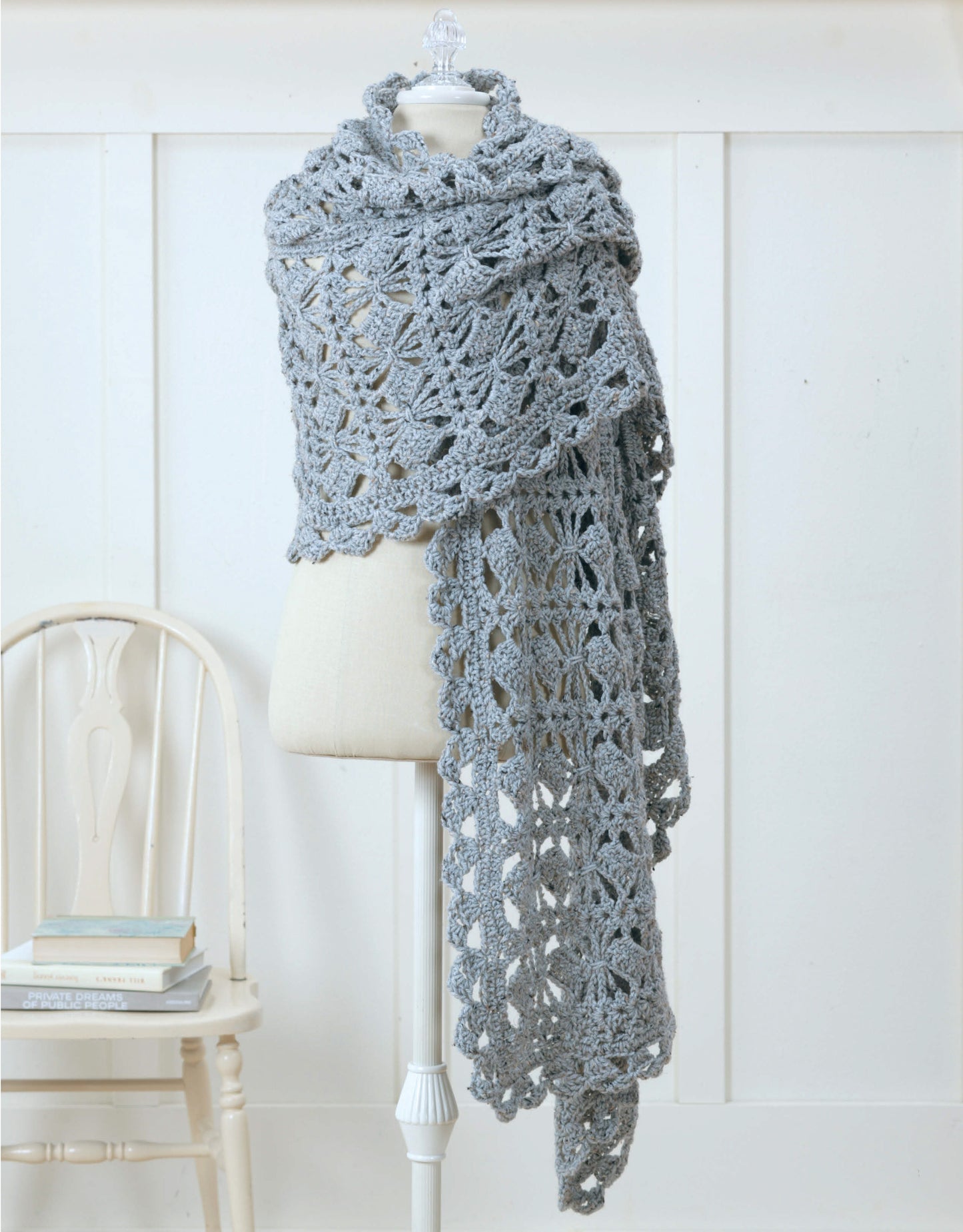 A chic light gray shawl from "Wraps by the Dozen" by Leisure Arts drapes elegantly over a dress form beside a white wooden chair and books, showcasing cozy style against a white paneled wall.