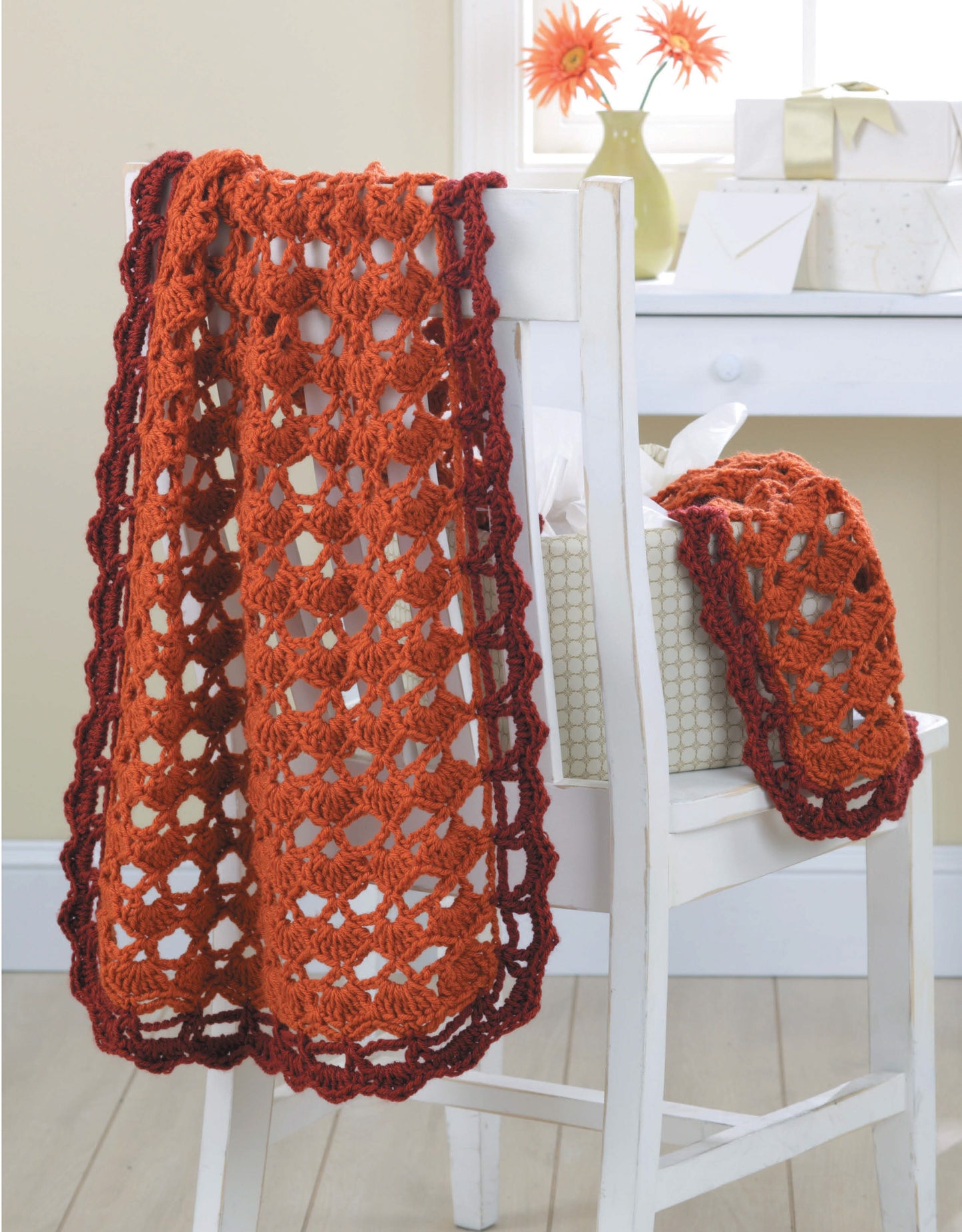 The Leisure Arts "Wraps by the Dozen" pattern features a red-orange crocheted lace wrap with a dark border draped over a white chair. A matching smaller wrap rests beside a gift box, and two orange flowers in a vase are on the windowsill.