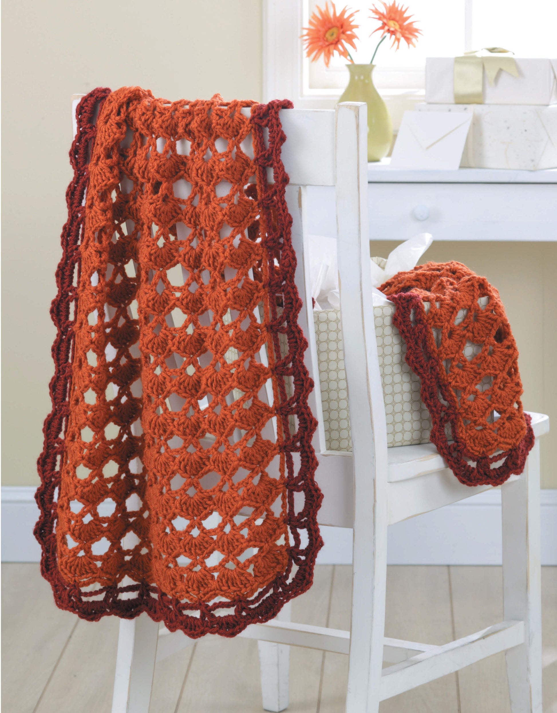 The Leisure Arts "Wraps by the Dozen" pattern features a red-orange crocheted lace wrap with a dark border draped over a white chair. A matching smaller wrap rests beside a gift box, and two orange flowers in a vase are on the windowsill.