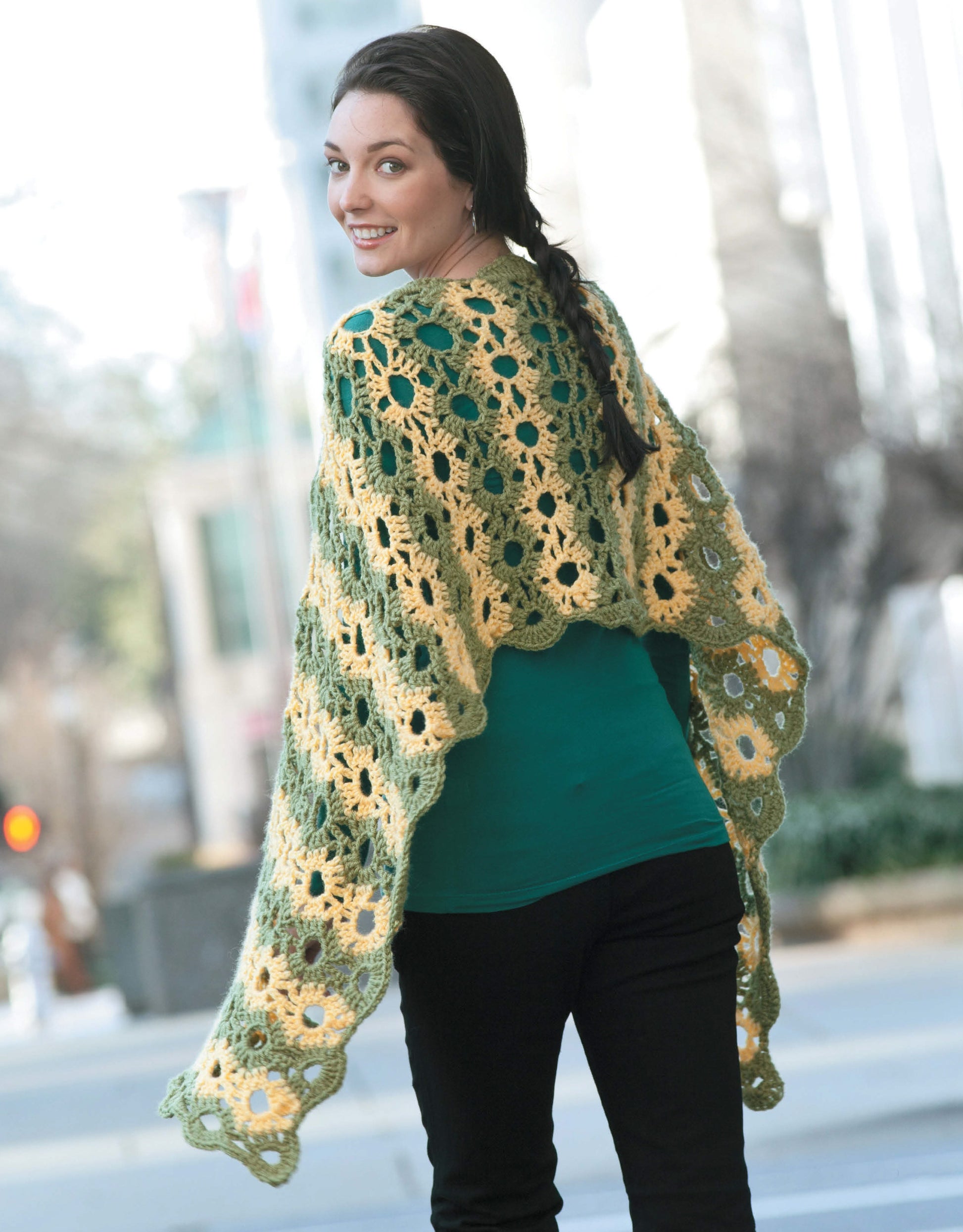 A woman outdoors smiles over her shoulder, modeling a yellow and green lacy shawl inspired by "Wraps by the Dozen—From Dressy to Casual, Show Your Style with Cozy, Fashionable Wraps" from Leisure Arts. Urban buildings and trees blur in the background.