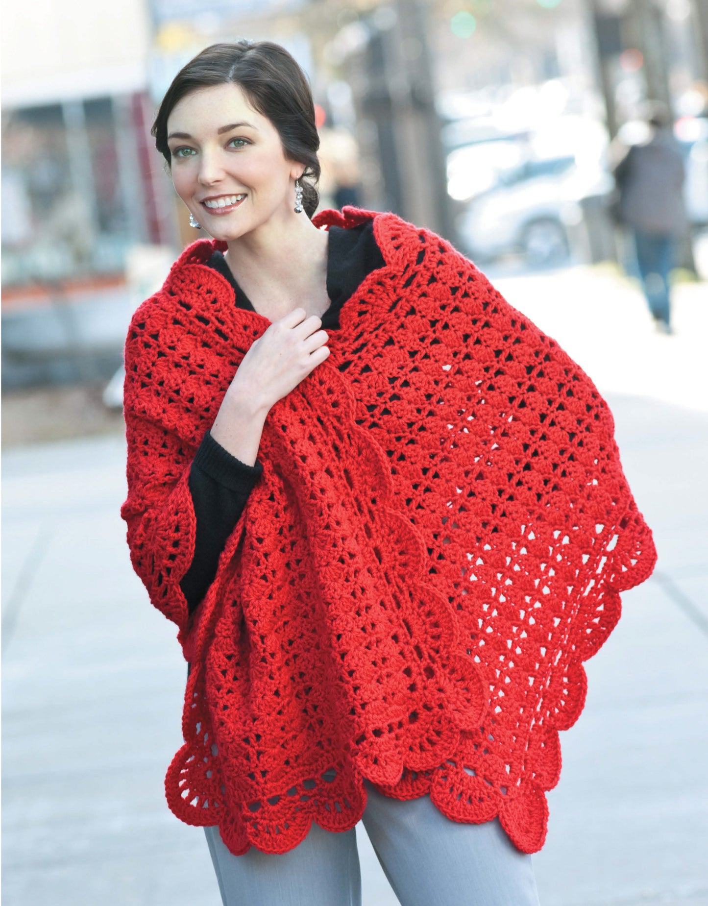 A woman smiles outdoors, wearing a bright red crochet shawl from "Wraps by the Dozen—From Dressy to Casual" by Leisure Arts—showcasing a beautiful, cozy worsted weight wrap perfect for stylish crochet enthusiasts.