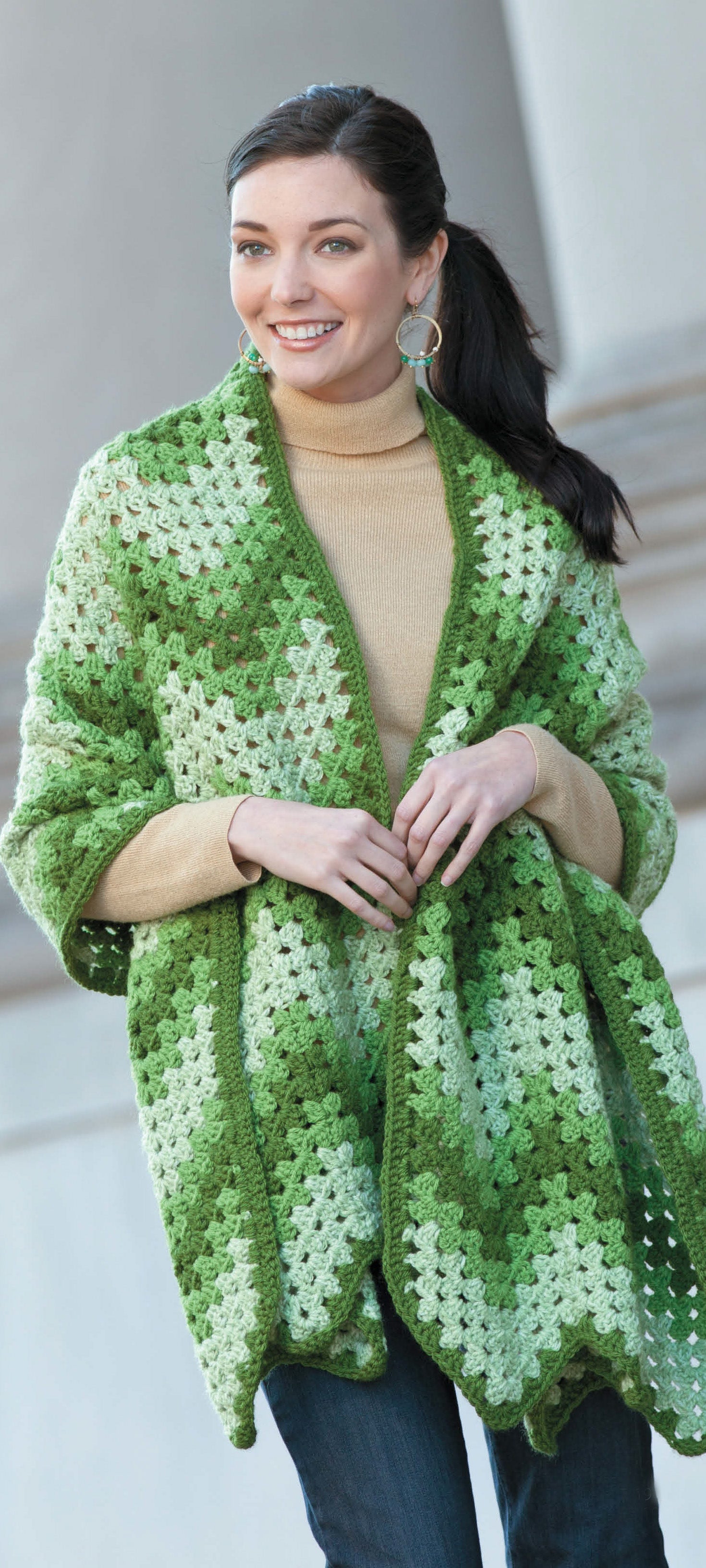 A woman with dark hair in a ponytail smiles while wearing a tan turtleneck, blue jeans, and a large green and white crochet wrap from "Wraps by the Dozen" by Leisure Arts—perfect cozy style for worsted weight yarn projects.