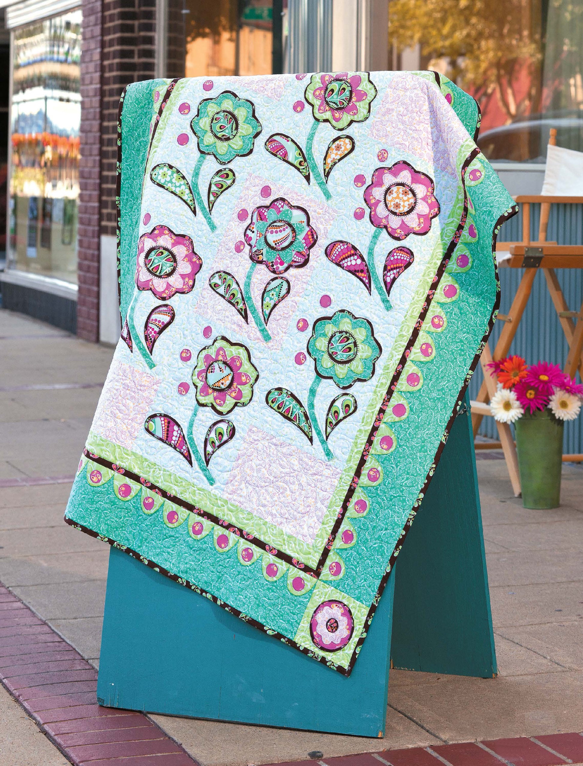 A quilt from Leisure Arts' City Walks—10 Contemporary Quilt Designs, with pink, green, and aqua floral motifs and borders, hangs over a teal stand on a sidewalk, with a street and shop windows in the background.