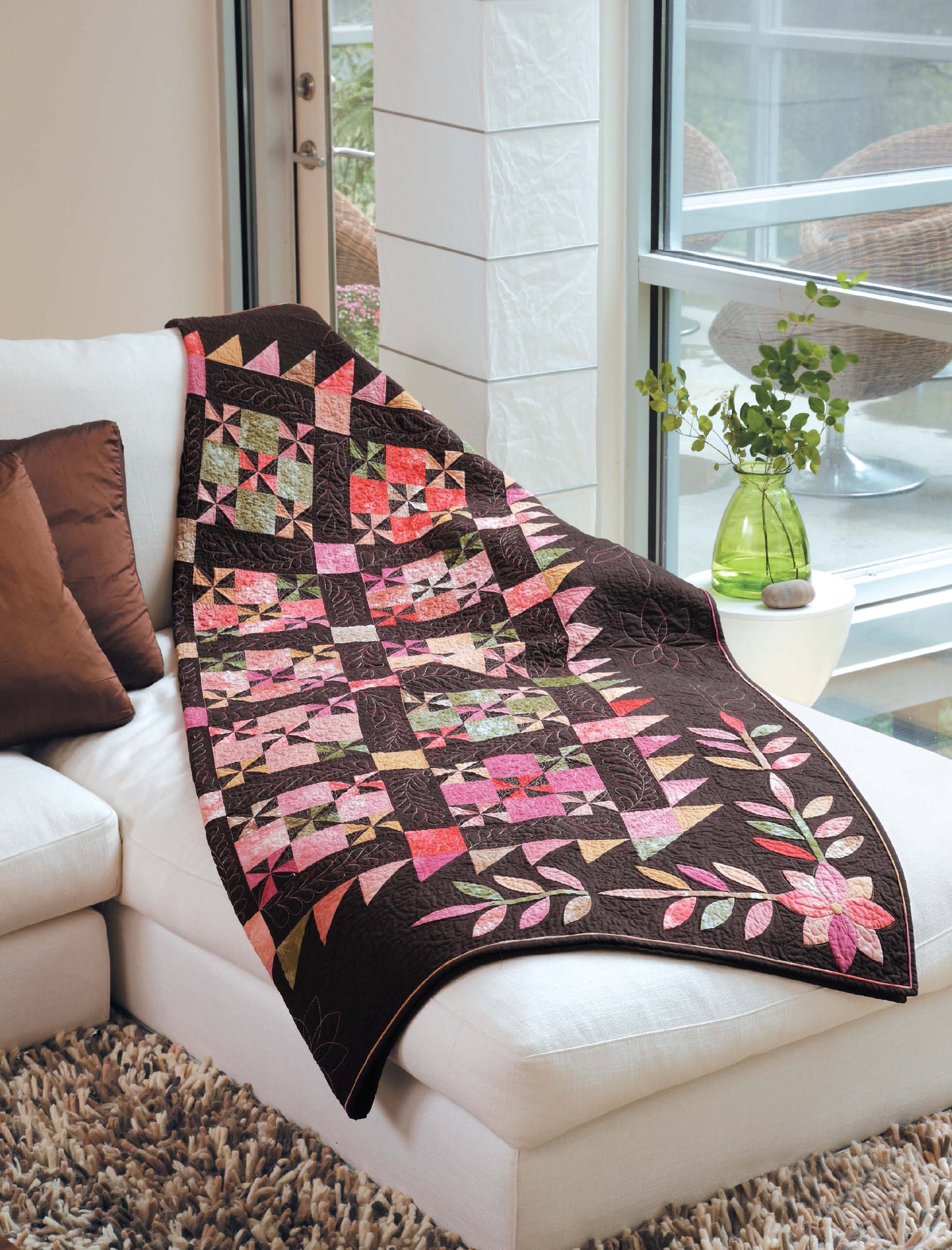 City Walks—10 Contemporary Quilt Designs by Leisure Arts adds vibrant style to a white sectional near sunlit windows, with a green vase nearby—ideal inspiration for intermediate quilting enthusiasts.